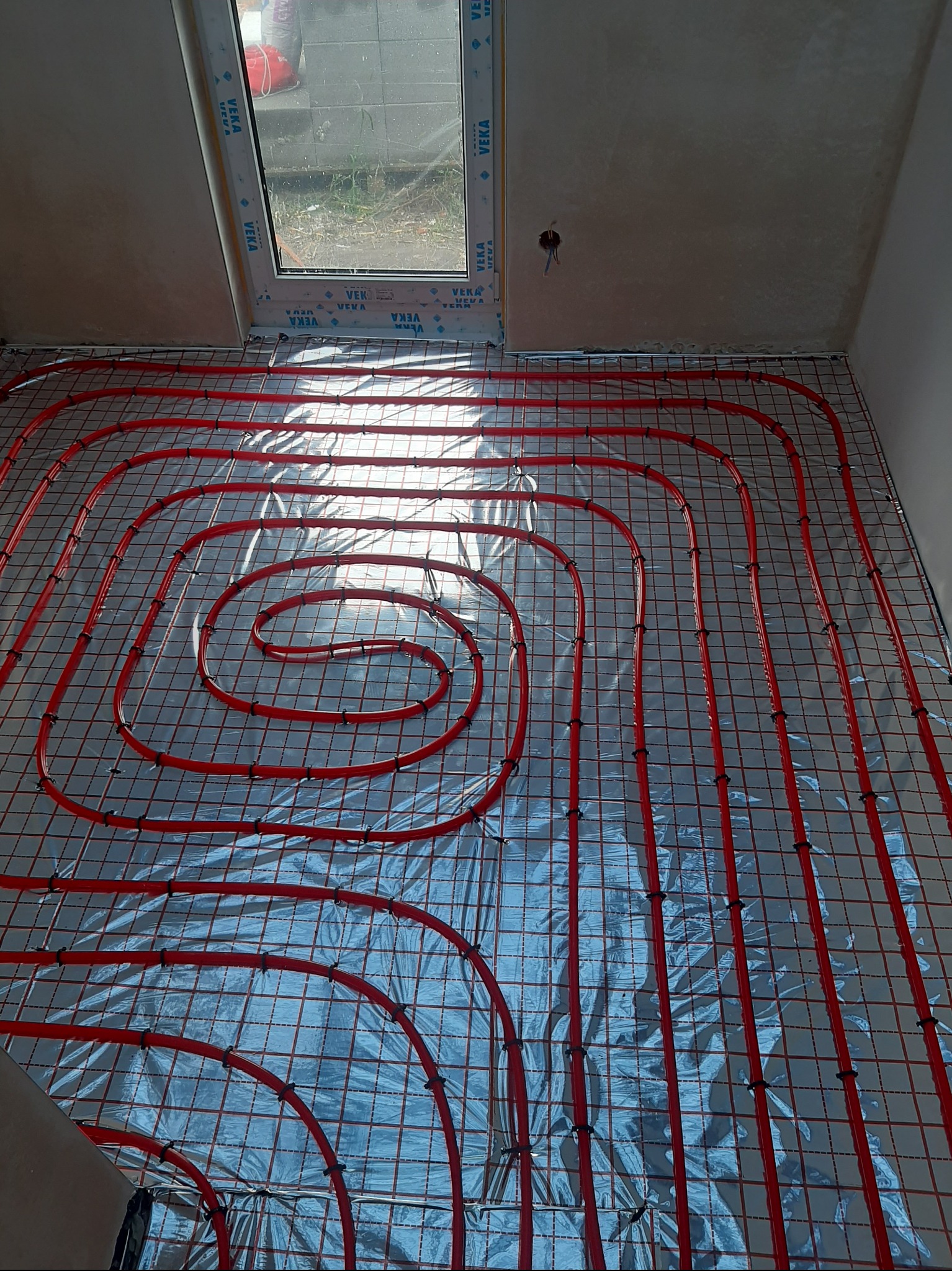 Czerwone rury ogrzewania podłogowego ułożone na srebrnej folii, widok z góry, w pomieszczeniu w Śremie. Rury przymocowane do siatki. Okno w tle.