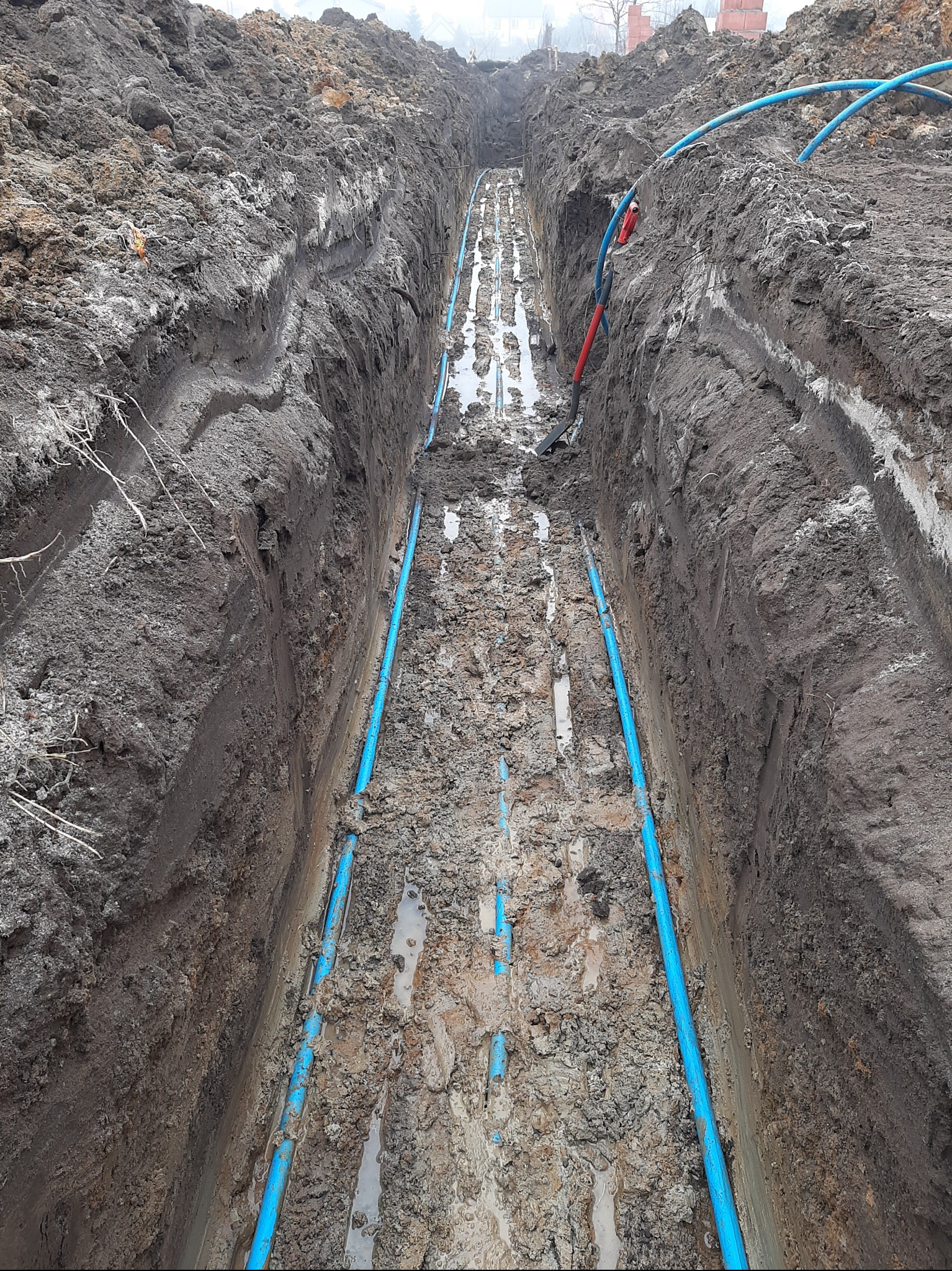 Wykop z ułożonymi niebieskimi rurami instalacji wodnej. W tle widoczny budynek. Na skraju wykopu leży szpadel z czerwoną rączką. Grunt mokry, gliniasty.