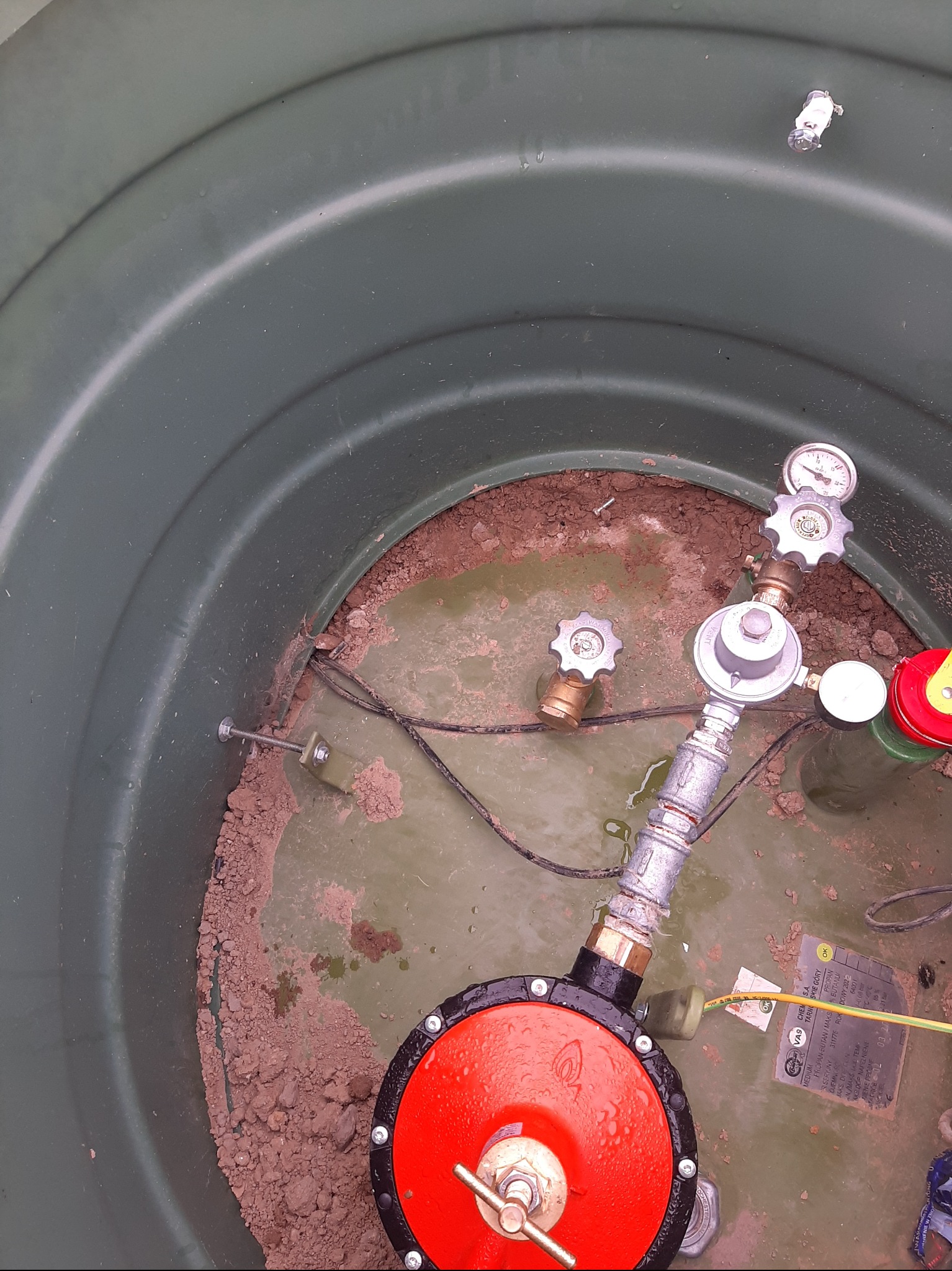 Wnętrze zbiornika z instalacją gazową: reduktor ciśnienia z manometrem, zawory i okablowanie. Widoczne elementy montażowe i ziemia na dnie zbiornika.