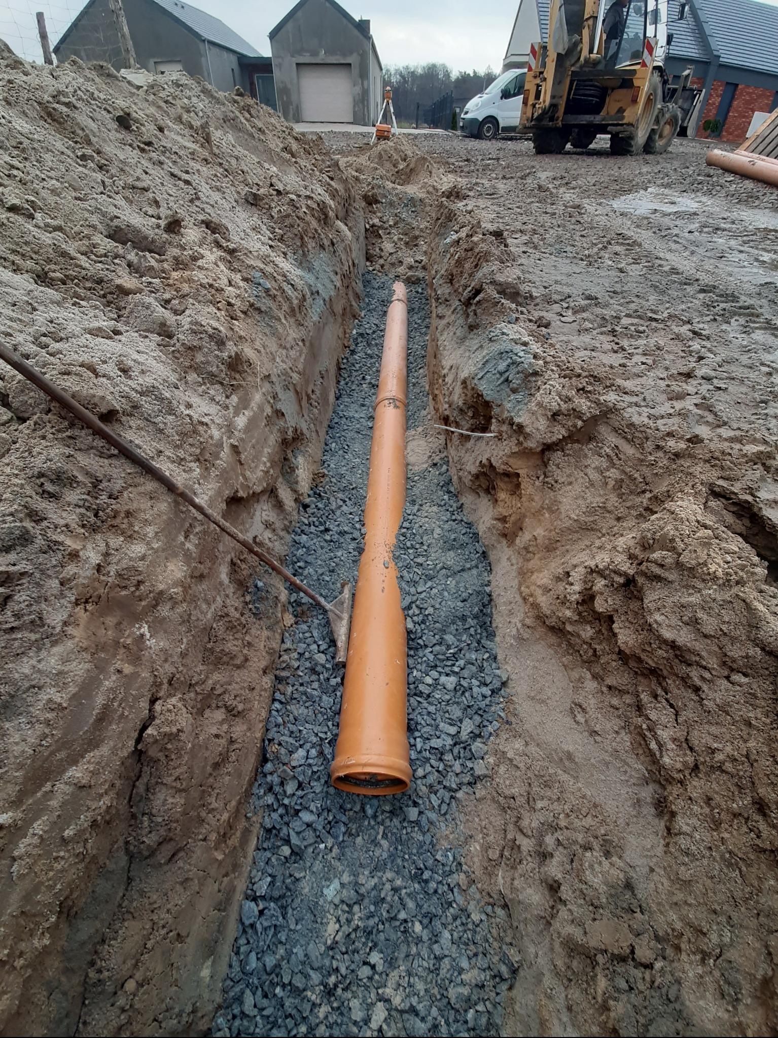 Pomarańczowa rura w wykopie wypełnionym żwirem, widok z góry. W tle budynki i sprzęt budowlany. Instalacja kanalizacyjna w trakcie realizacji.