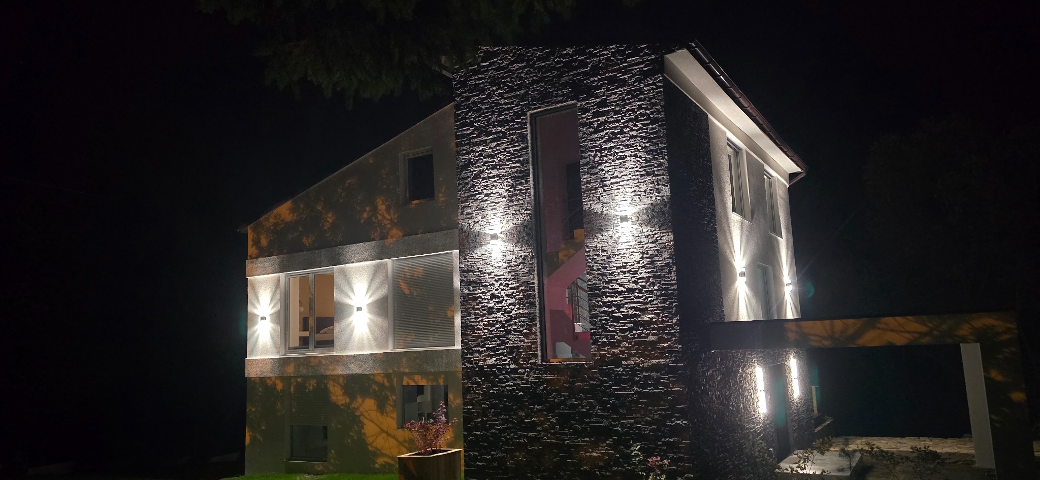Nowoczesny dom dwukondygnacyjny z elewacją z kamienia naturalnego, oświetlony nocą ciepłym światłem. Widoczne okna z roletami i fragment roślinności w donicy.