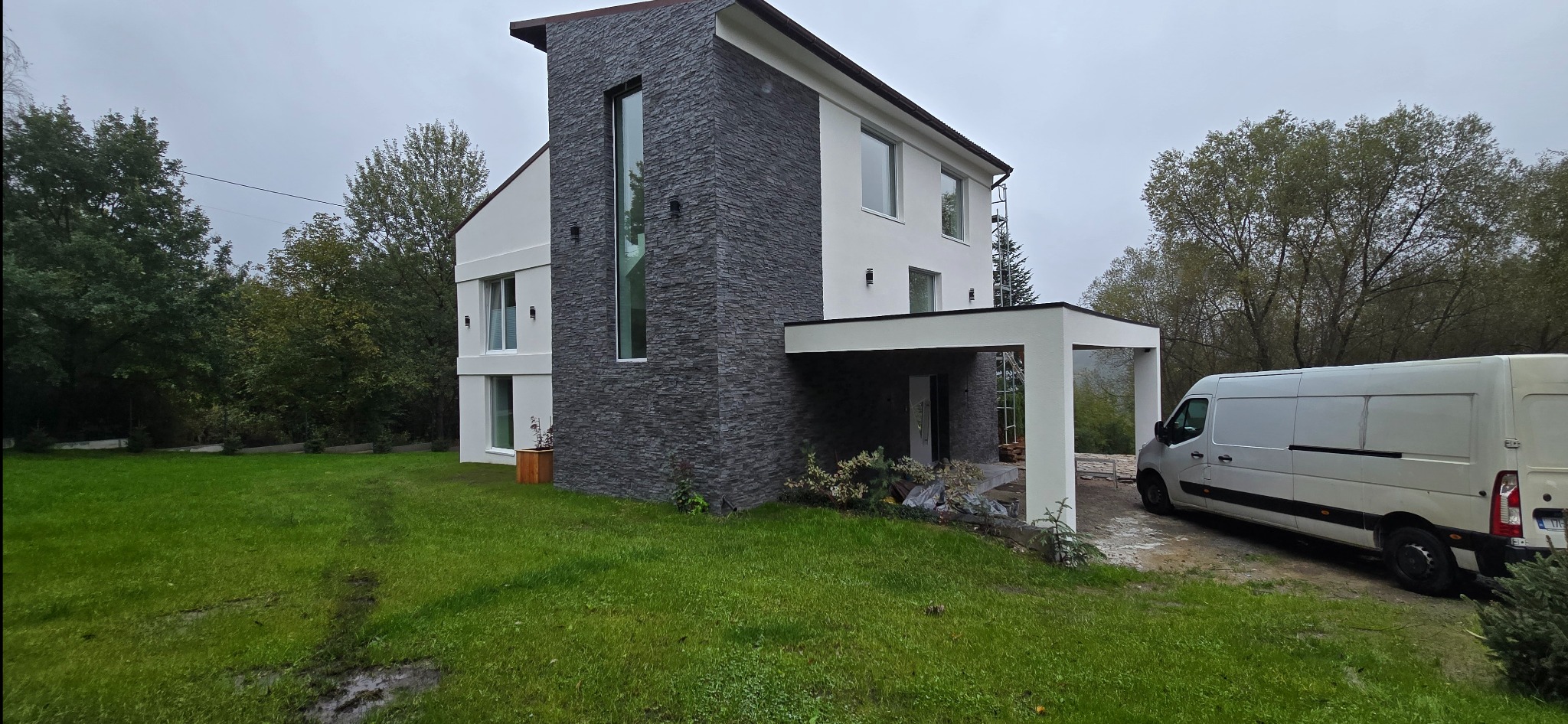 Nowoczesny dom z elewacją z szarego kamienia i białym tynkiem, widoczny z trawnika. Na podjeździe stoi biały bus. W tle drzewa i pochmurne niebo.