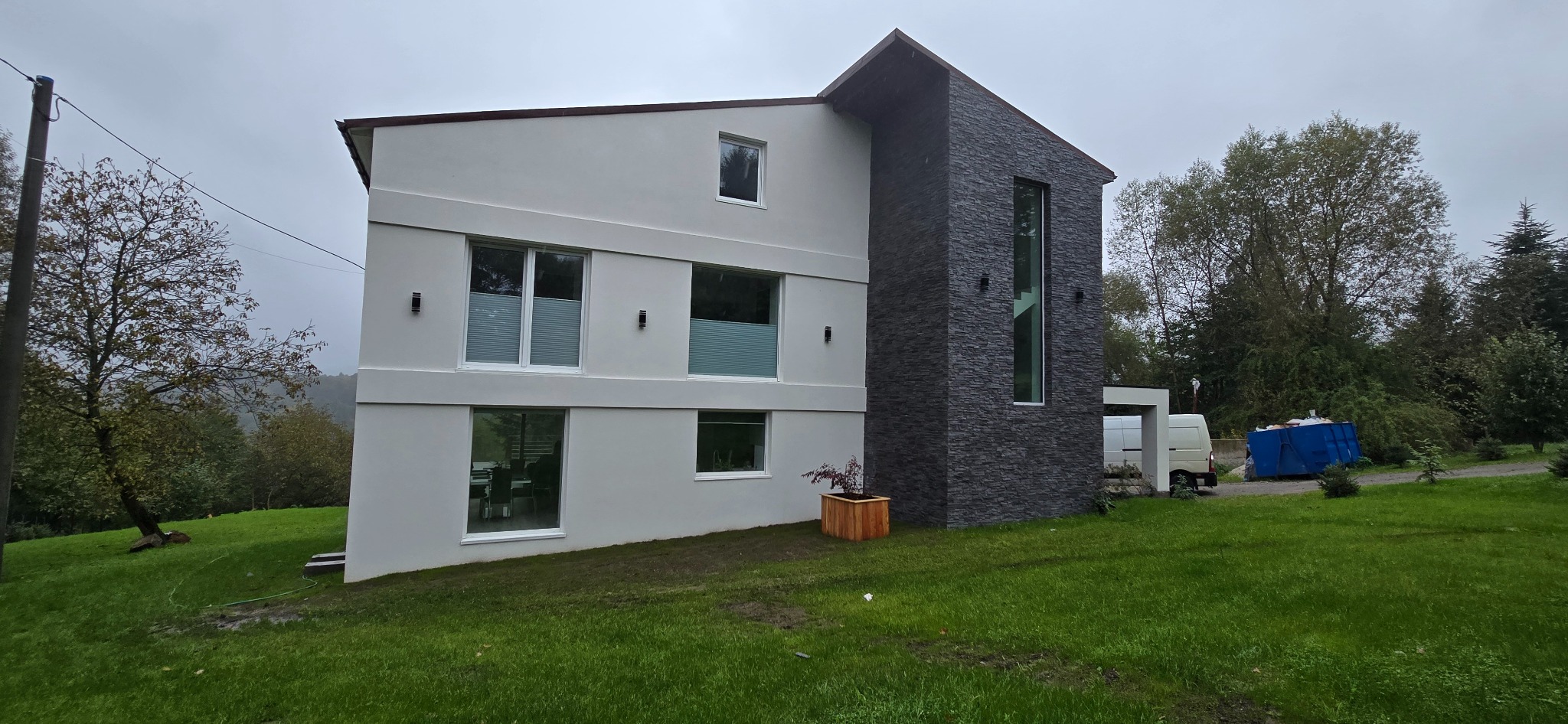 Elewacja dwukondygnacyjnego domu w Mucharzu z jasnym tynkiem i ciemną okładziną kamienną. Widoczne okna z roletami i oświetlenie zewnętrzne. Na trawniku skrzynia z rośliną.