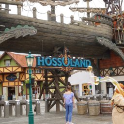 ARTISAN ALEKS COMPANY - Wejście do Hossoland: drewniana konstrukcja stylizowana na statek, brama z napisem, budynki w tle, osoba puszczająca bańki mydlane, barierki wejściowe.