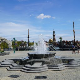 ARTISAN ALEKS COMPANY - Fontanna z szarego kamienia z tryskającą wodą w Rumi. W tle budowle stylizowane na skandynawskie. Dzień słoneczny, błękitne niebo z chmurami.