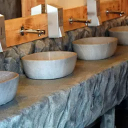 Kamienne umywalki w rzędzie, z nowoczesną armaturą i dozownikami. Umywalki w kształcie mis, osadzone na blacie imitującym skałę. Drewniane elementy wykończenia.
