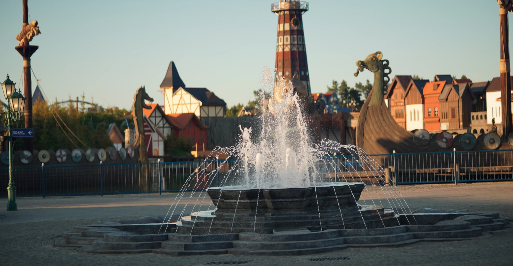 Fontanna w parku rozrywki w Rumi, na tle stylizowanych budynków i statku wikingów. Ujęcie z bliska, woda tryska w górę, tworząc efektowny widok.