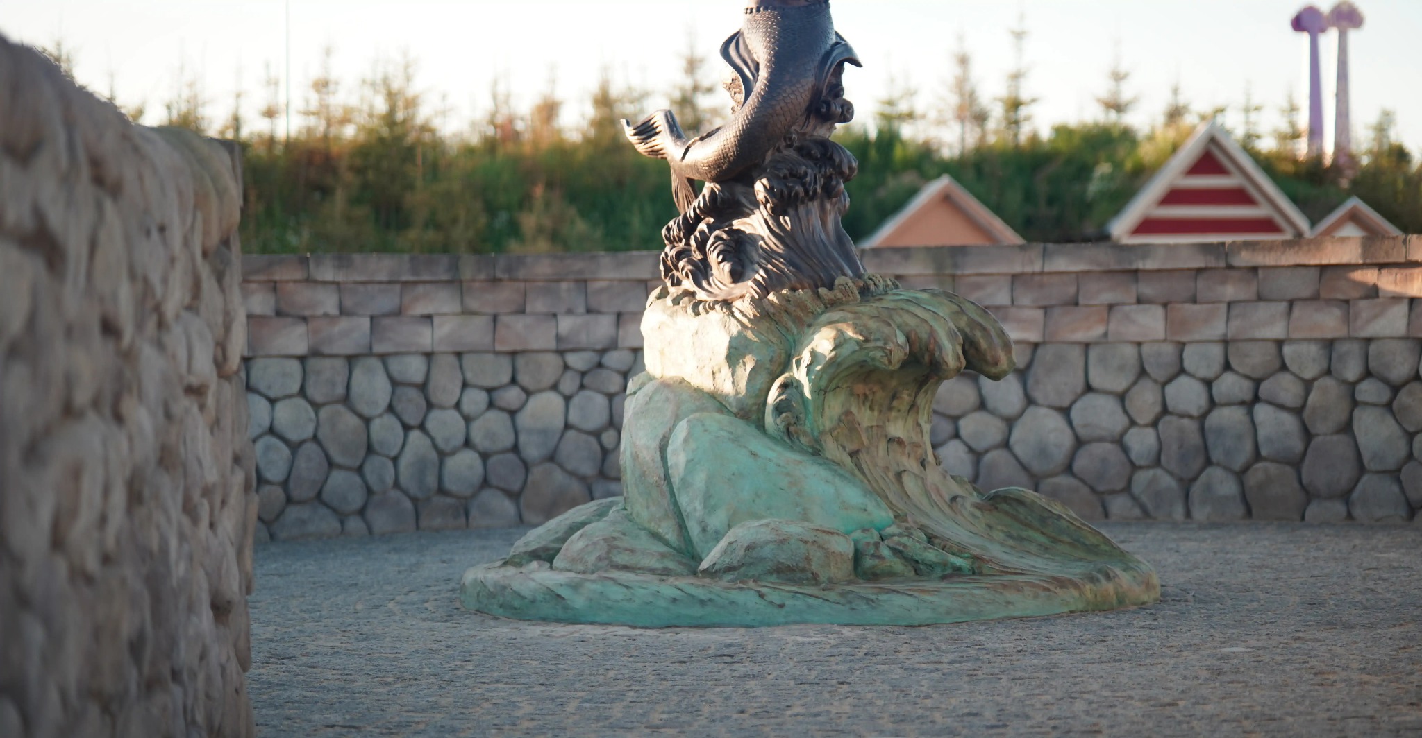 Fontanna z rzeźbą syreny na tle kamiennej ściany i zieleni. Dekoracja ogrodowa. Ujęcie z dołu podkreśla detale rzeźby i otoczenia.