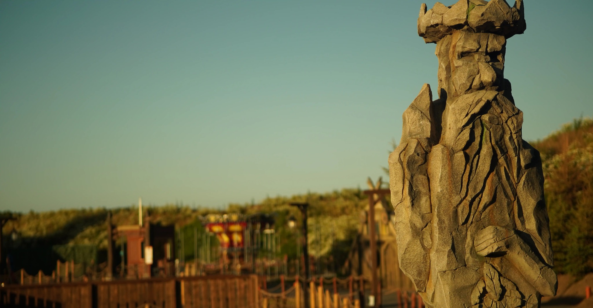 Kamienna rzeźba króla z koroną na tle placu zabaw i zieleni. Delikatne oświetlenie podkreśla fakturę kamienia. Detal z bliska ukazuje rzemiosło artystyczne.