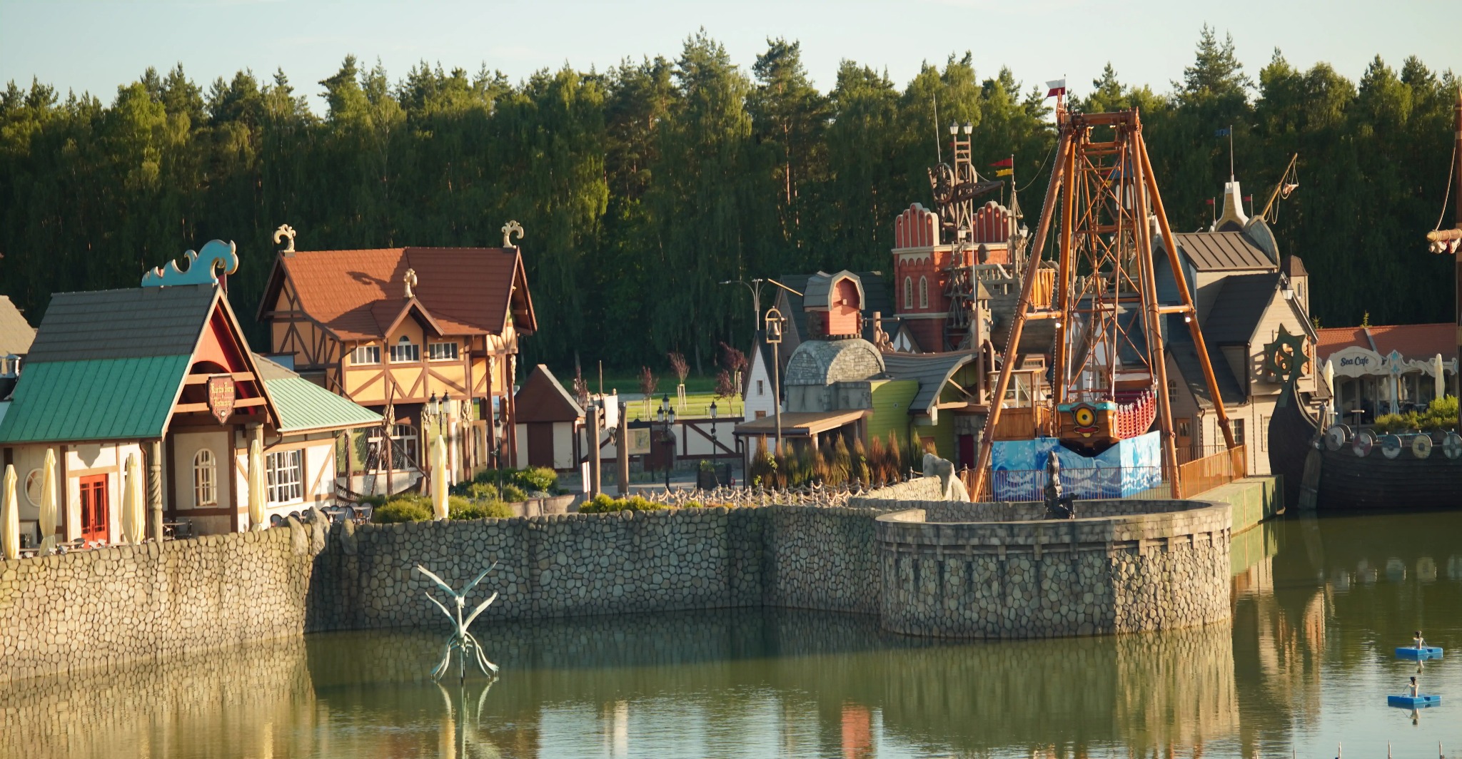 Bajkowa zabudowa nad wodą z elementami architektury średniowiecznej i parkiem rozrywki w tle. Widok na kamienną zabudowę i rzeźbę na wodzie.