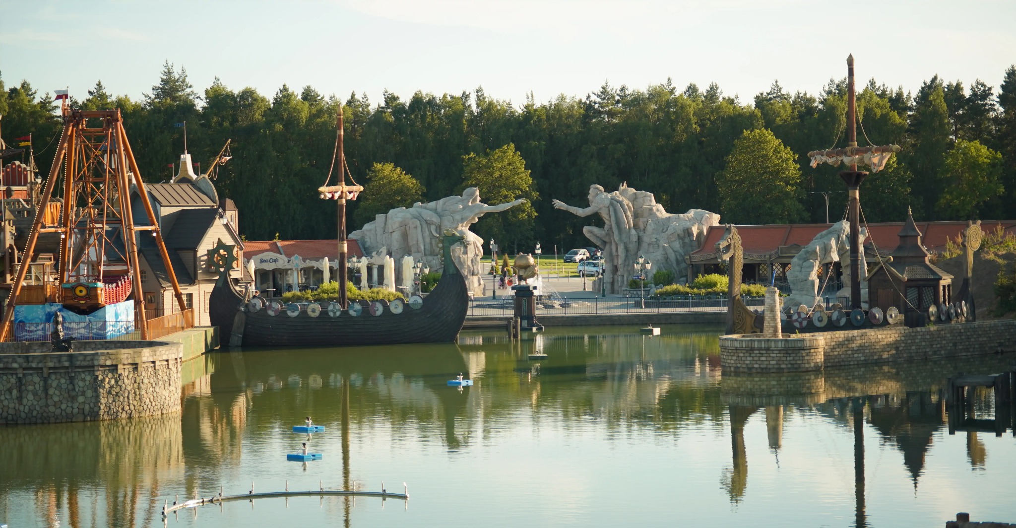 Park rozrywki z replikami łodzi Wikingów, rzeźbami i budynkami o tematyce nordyckiej, odbijającymi się w spokojnej wodzie jeziora.