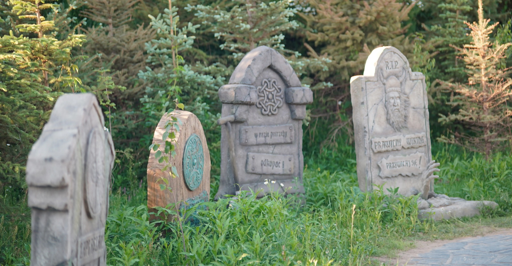 Rząd fantazyjnych nagrobków z napisami i zdobieniami, umieszczonych w zielonym otoczeniu przypominającym leśną scenerię. Jeden z nich z wizerunkiem Wikinga.