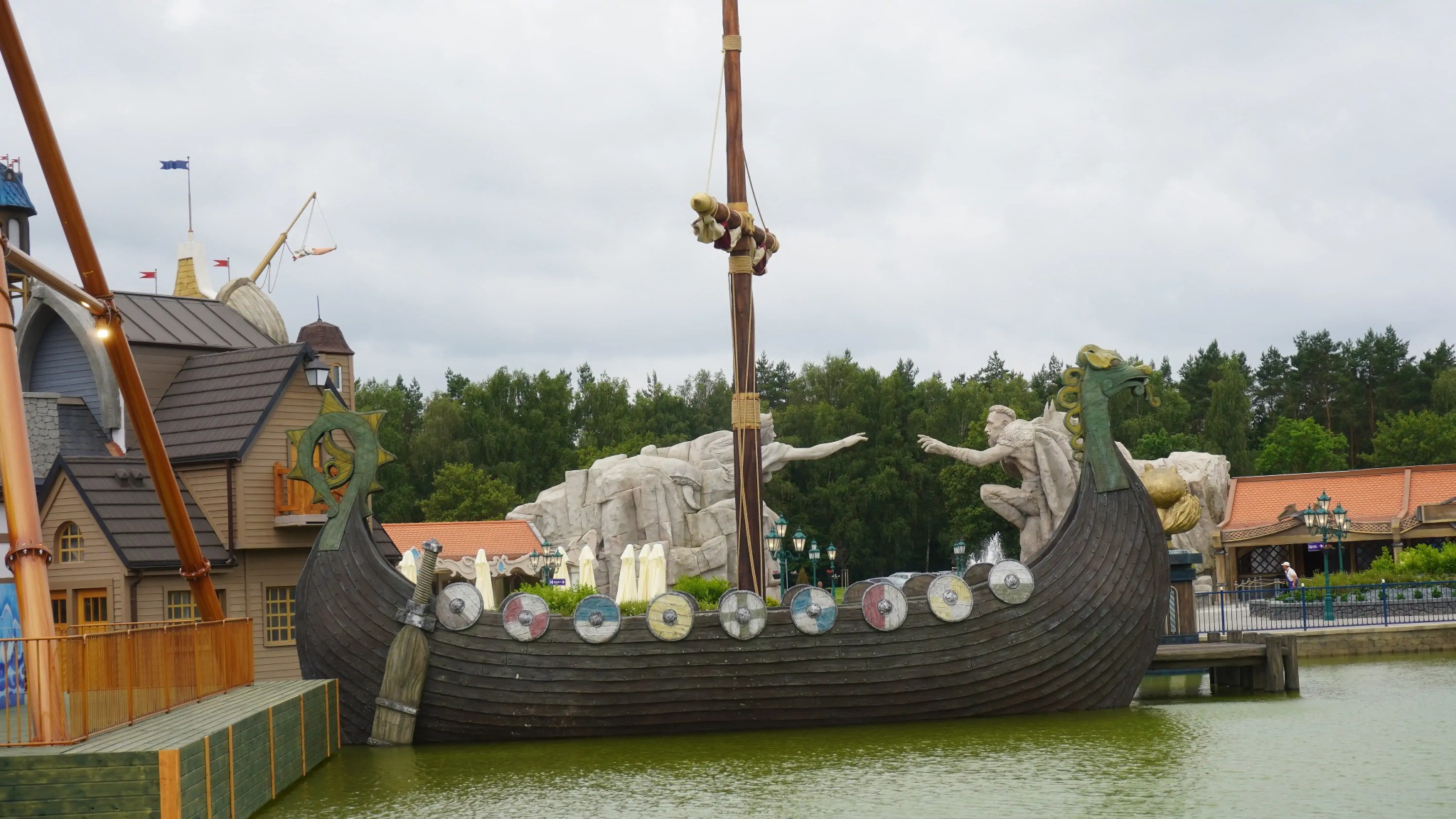 Replika łodzi Wikingów na wodzie, z rzeźbami i budynkami w tle, tworząca scenografię parku rozrywki o tematyce historycznej.