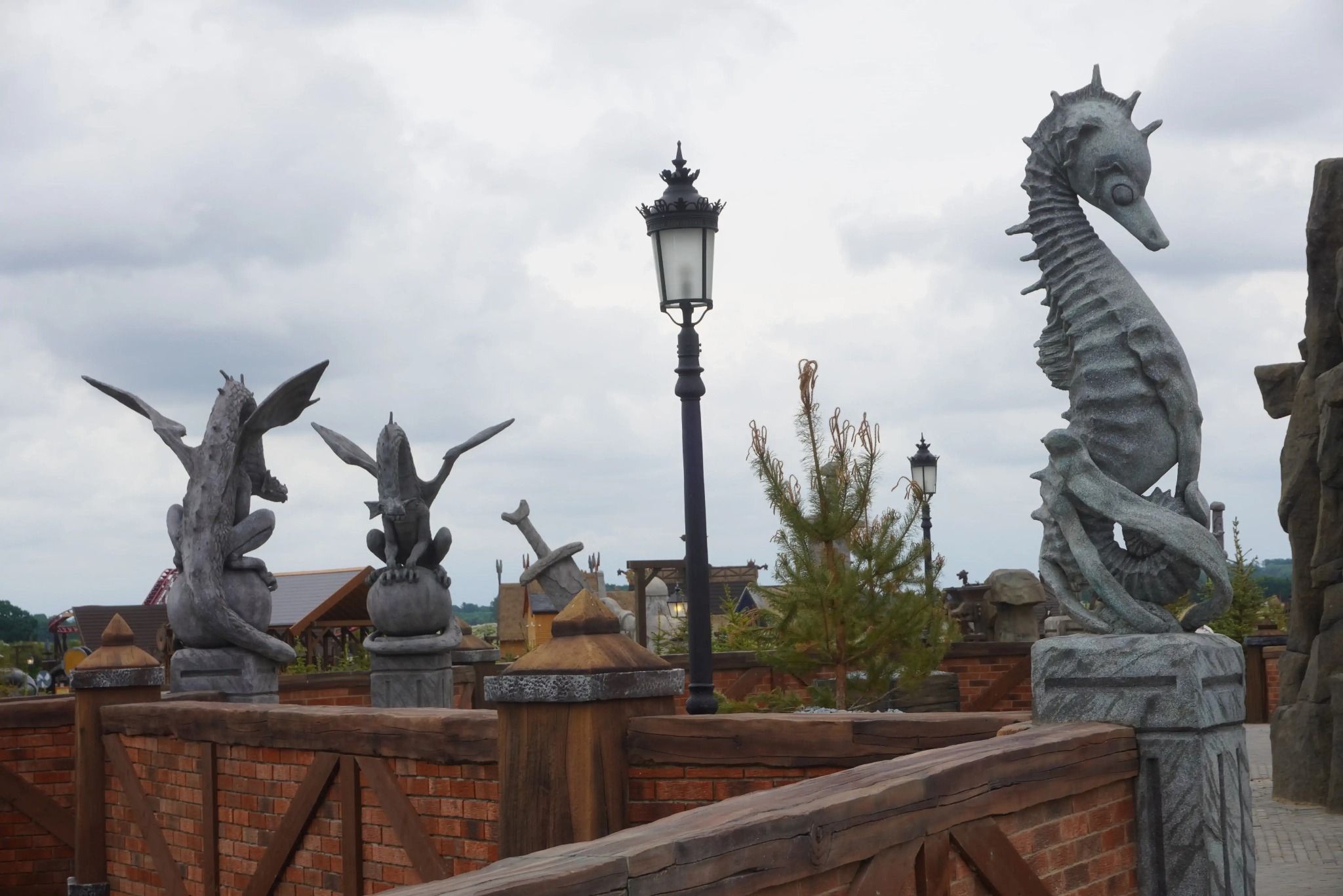 Fantazyjny park tematyczny: rzeźby smoków i konika morskiego na ceglanym murze, oświetlone latarnią, pod pochmurnym niebem. Wyjątkowa aranżacja przestrzeni.