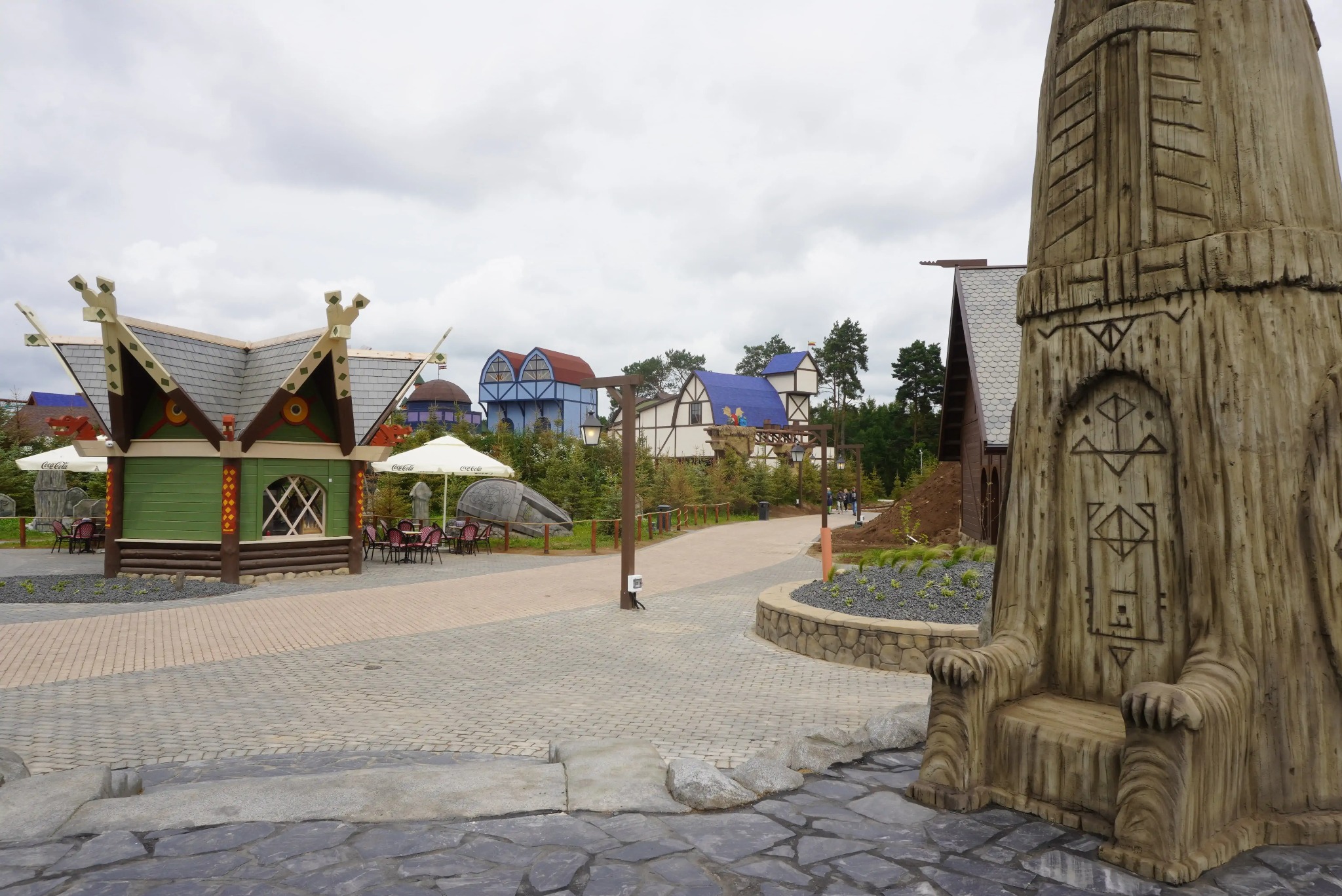 Tematyczny park rozrywki: kolorowe budynki o fantazyjnej architekturze, brukowane ścieżki i rzeźba tronu z drewna. Widok na otwartą przestrzeń w pochmurny dzień.