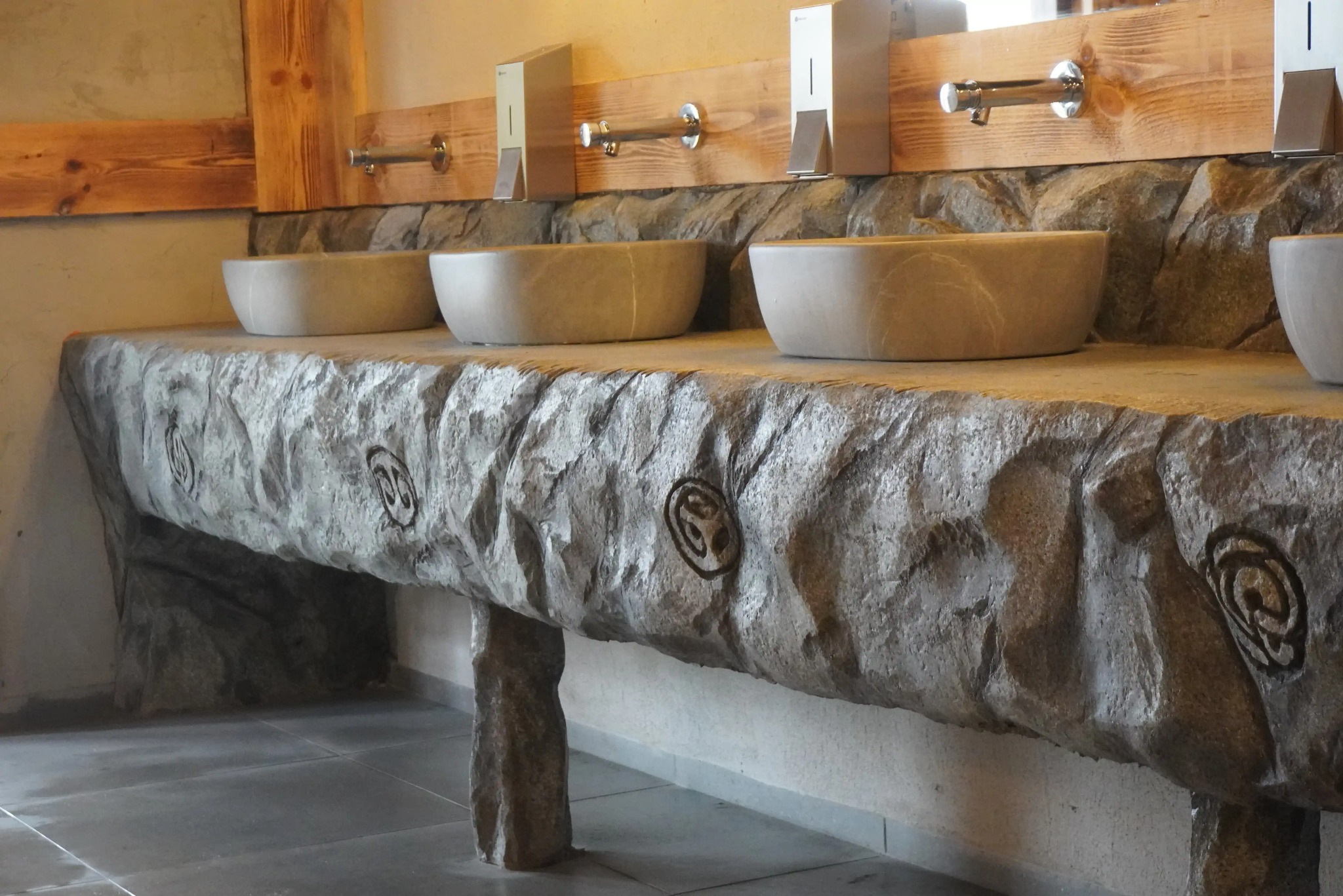 Kamienna umywalka z trzema misami i kranami, drewniane wykończenie nad blatem. Dekoracyjny wzór na kamieniu. Wykończenie łazienki w stylu rustykalnym.