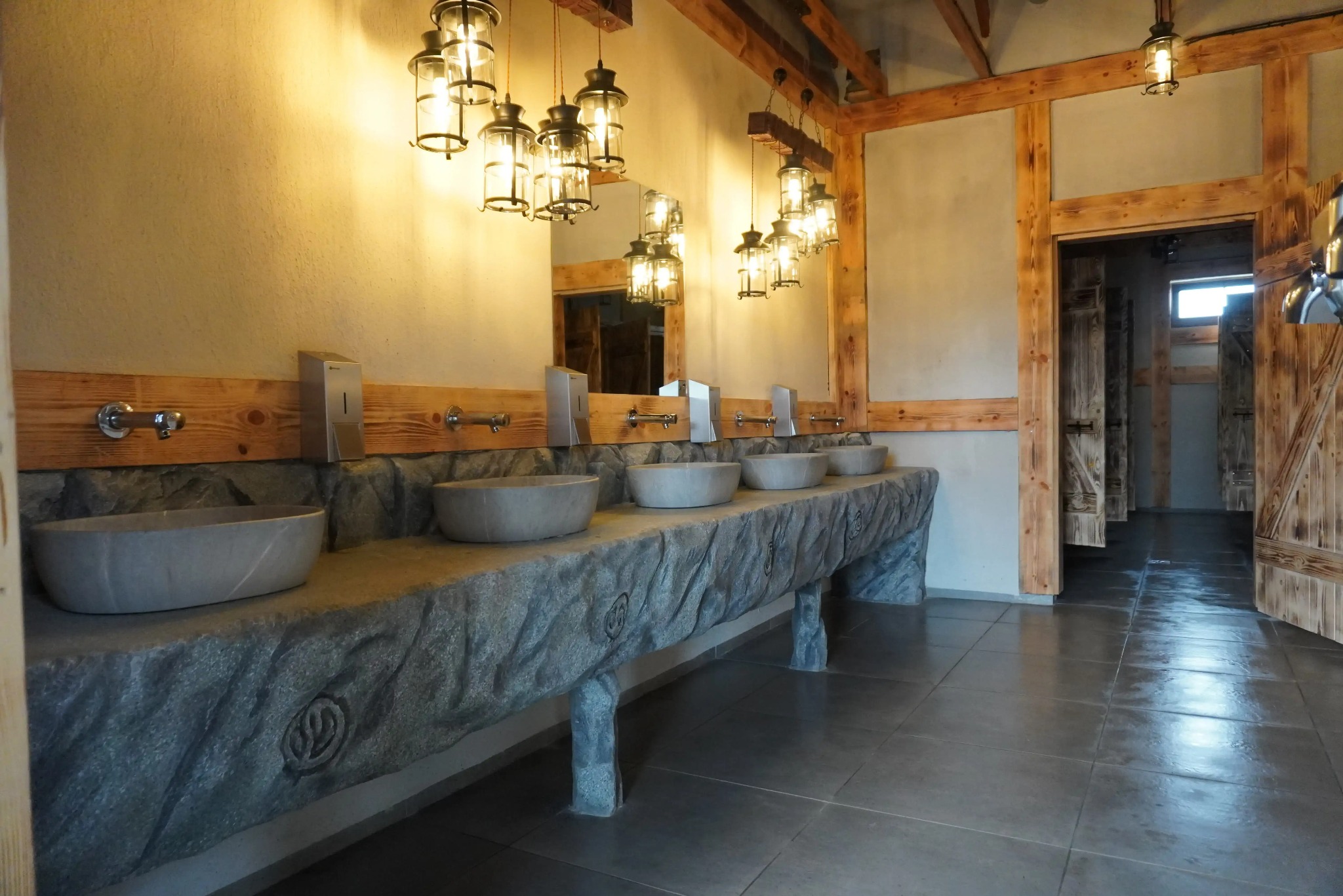 Rustykalna łazienka publiczna z kamienną umywalką, drewnianymi elementami i wiszącymi lampami w stylu retro. W tle widoczne kabiny z drewnianymi drzwiami.