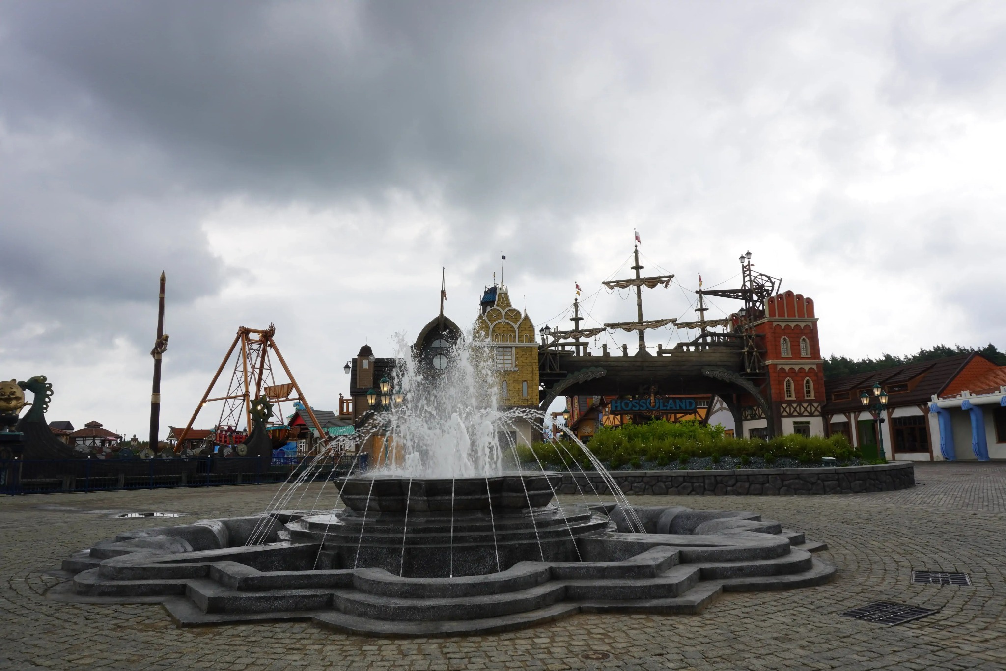 Fontanna w parku rozrywki z budynkami w stylu pirackim i karuzelami w tle, pod pochmurnym niebem. Detale architektoniczne i tryskająca woda.