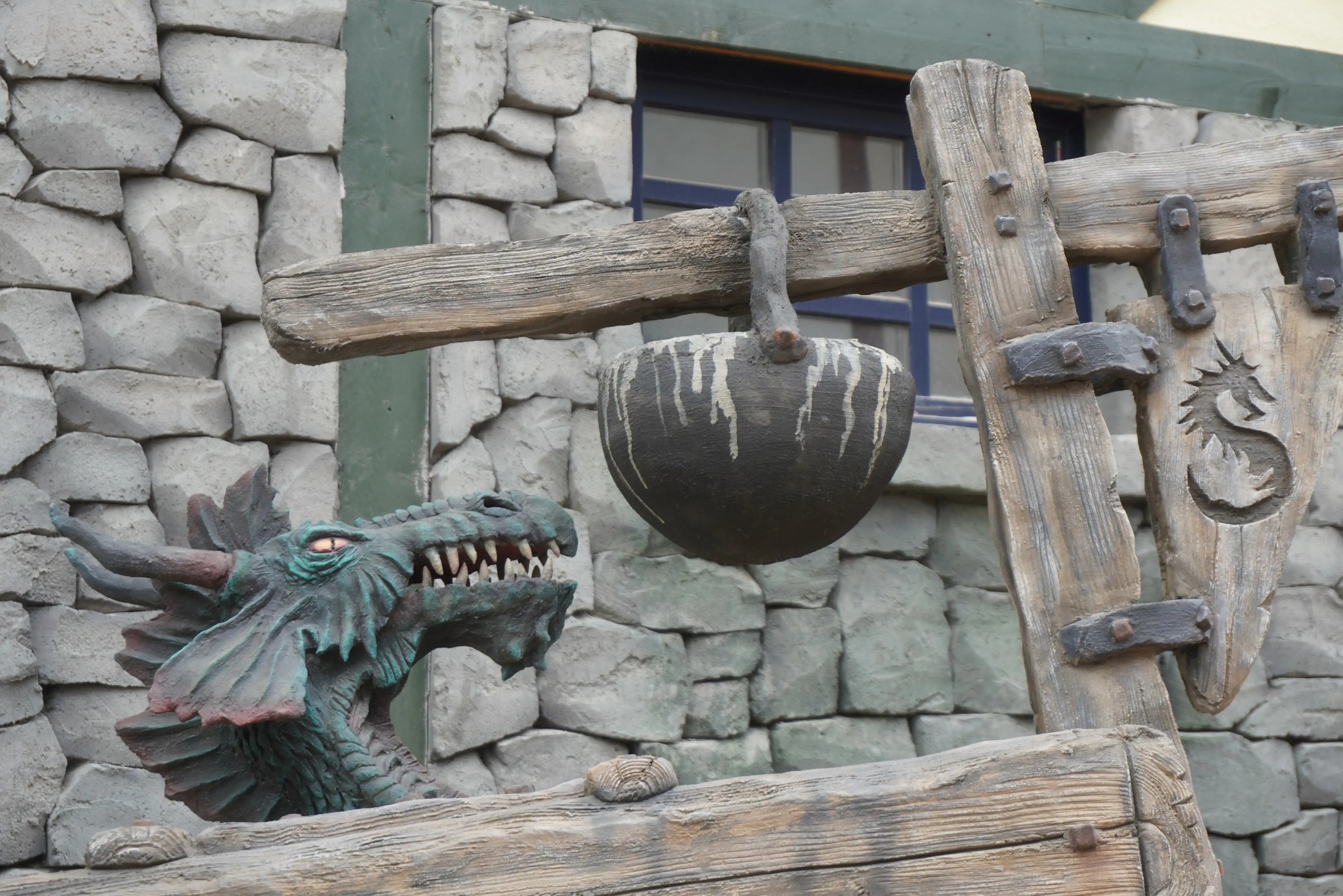Dekoracyjna ściana z kamienia z rzeźbą smoka i drewnianą konstrukcją z kociołkiem, stylizowana na średniowieczny zamek, okno w tle.