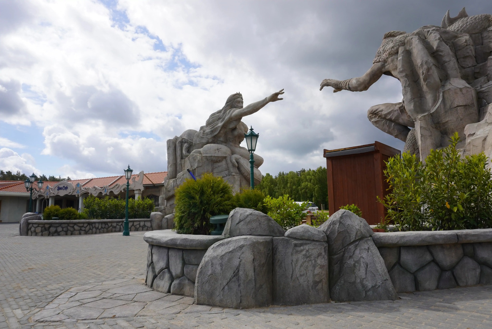 Park z rzeźbami w szarym kamieniu, imitującymi biblijną scenę stworzenia Adama. W tle budynek Sea Cafe i dekoracyjne lampy parkowe.