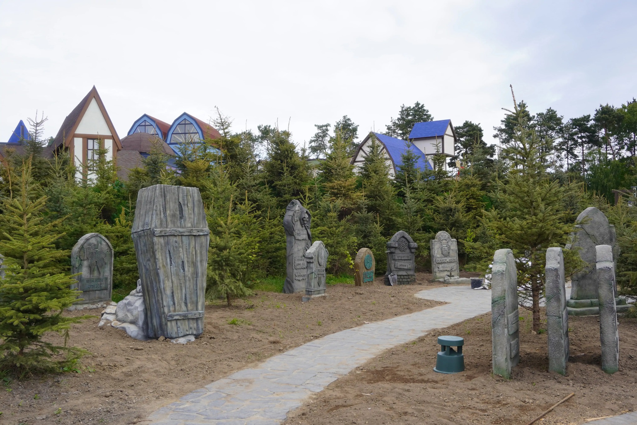 Tematyczny park rozrywki: ścieżka z nagrobkami i trumną, w tle budynki z kolorowymi dachami, otoczone zielenią iglaków. Nietypowy krajobraz.