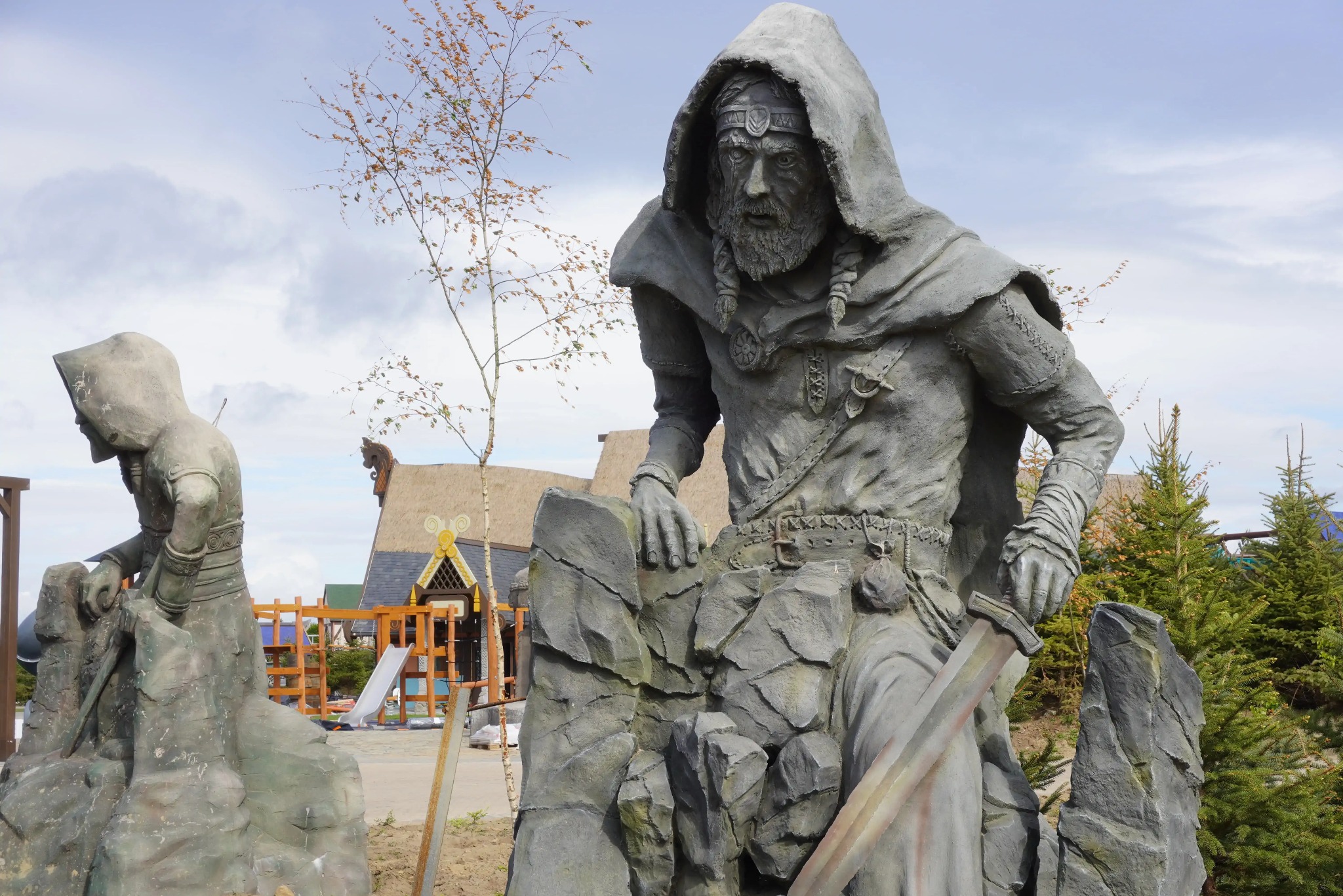 Dwie szare, kamienne rzeźby w parku: jedna przedstawia postać w kapturze z mieczem, druga skuloną postać. W tle plac zabaw i drzewa.