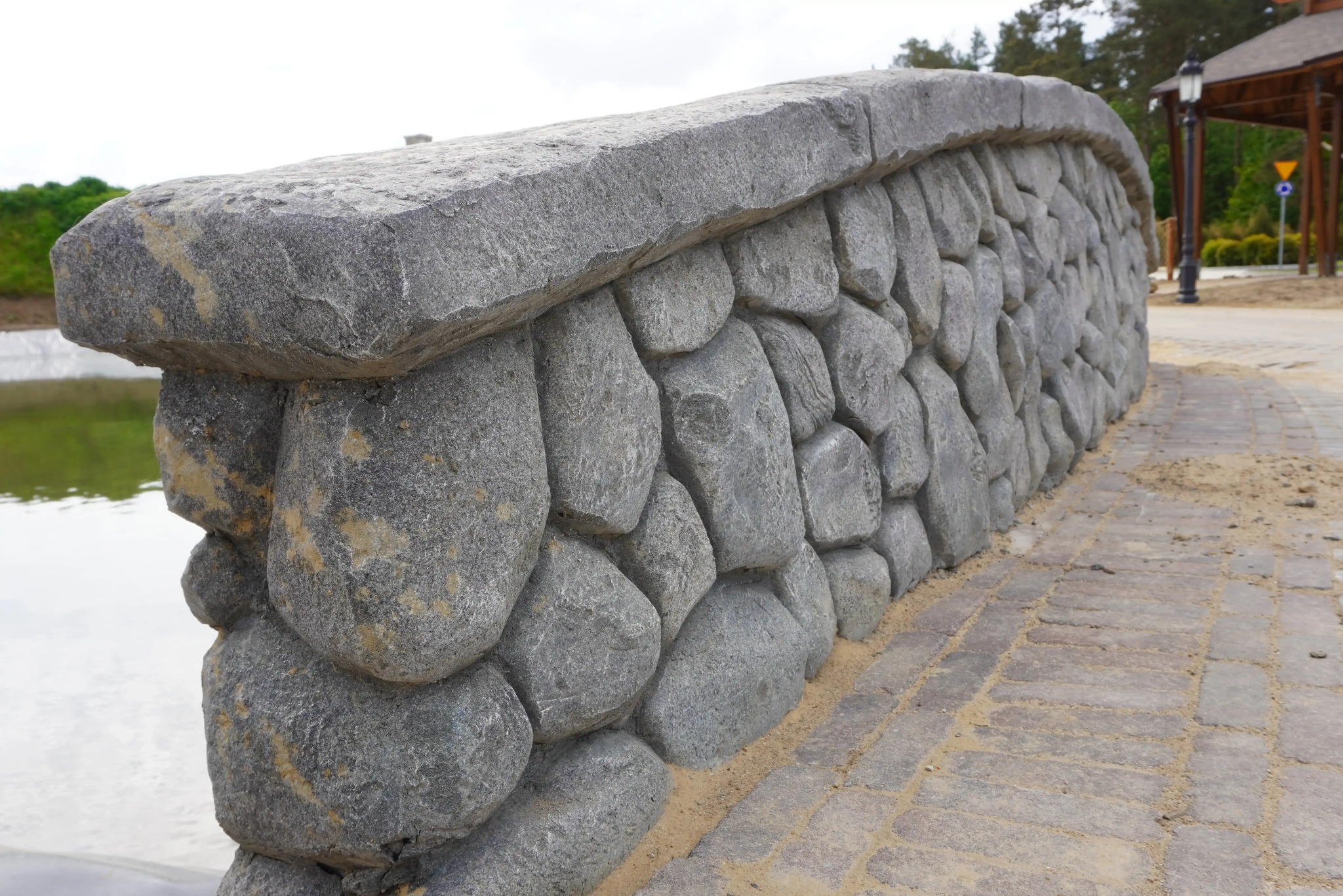 Kamienny murek z okrąglaków, zwieńczony płaskim kamieniem, tworzący barierę przy ścieżce. W tle woda i zieleń. Widoczne detale kamieni i faktura muru.
