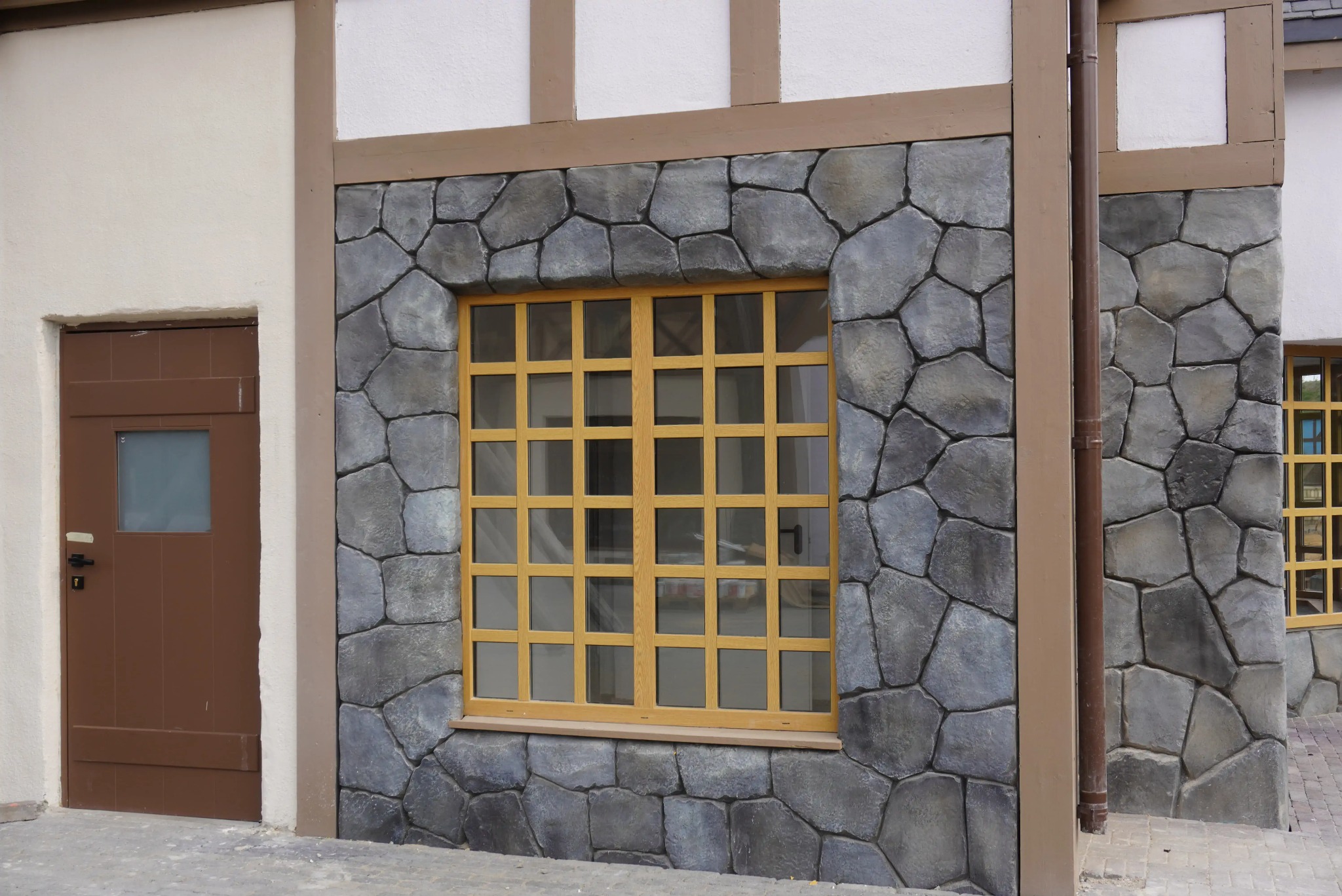 Elewacja budynku z oknem w żółtej ramie i drzwiami. Kamienna okładzina, imitacja muru. Detal architektoniczny, stylizacja.