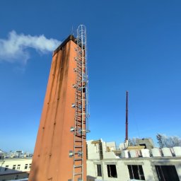 Wysoki, pomarańczowy komin z metalową drabiną na dachu budynku w zimowej scenerii, na tle błękitnego nieba. Widoczne ślady zabrudzeń na kominie.