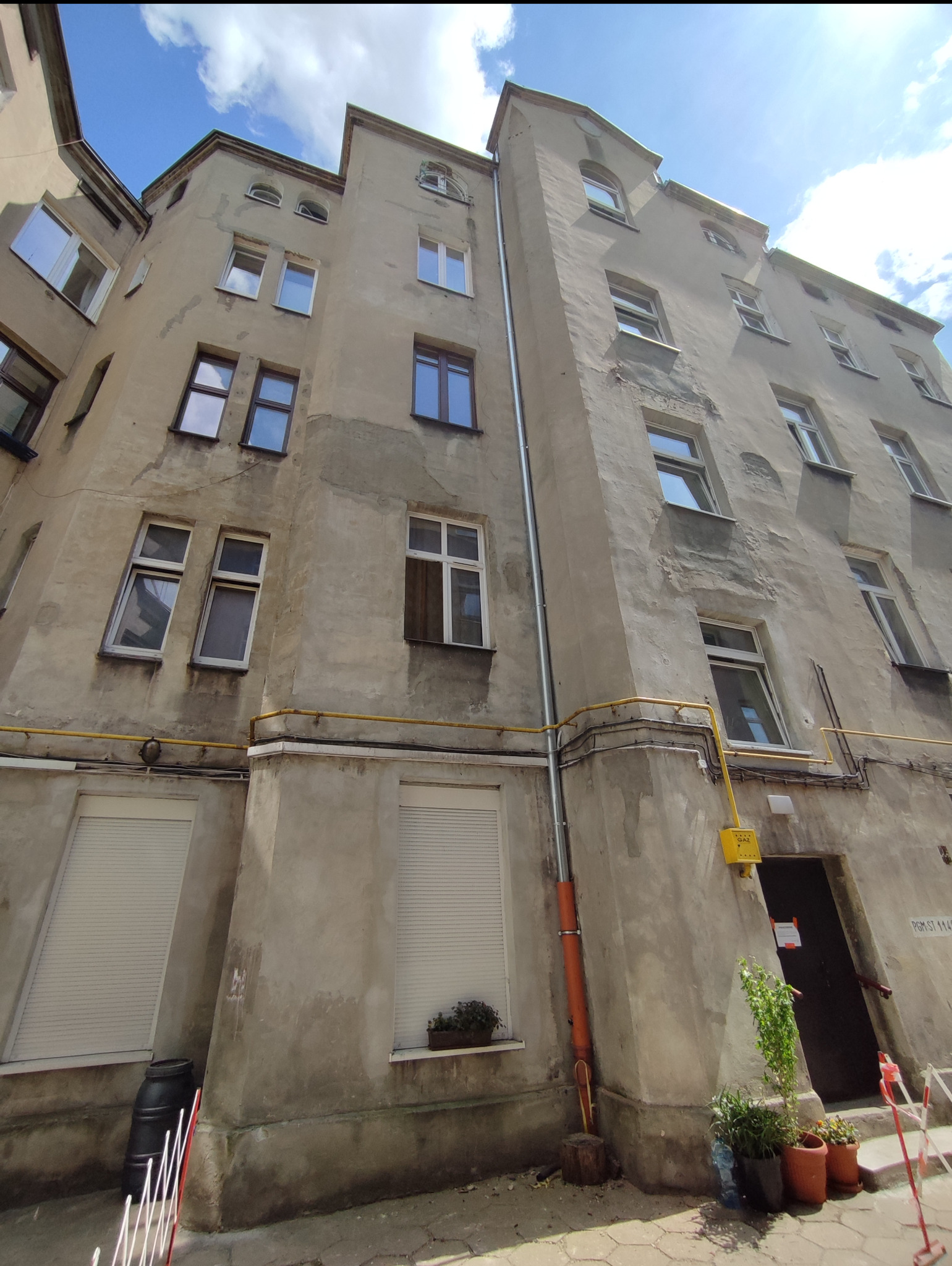 Kamienica z nową, pionową rurą spustową, widoczna z dołu pod kątem. Szara elewacja, okna z roletami, żółta instalacja gazowa i rośliny w donicach przy wejściu.