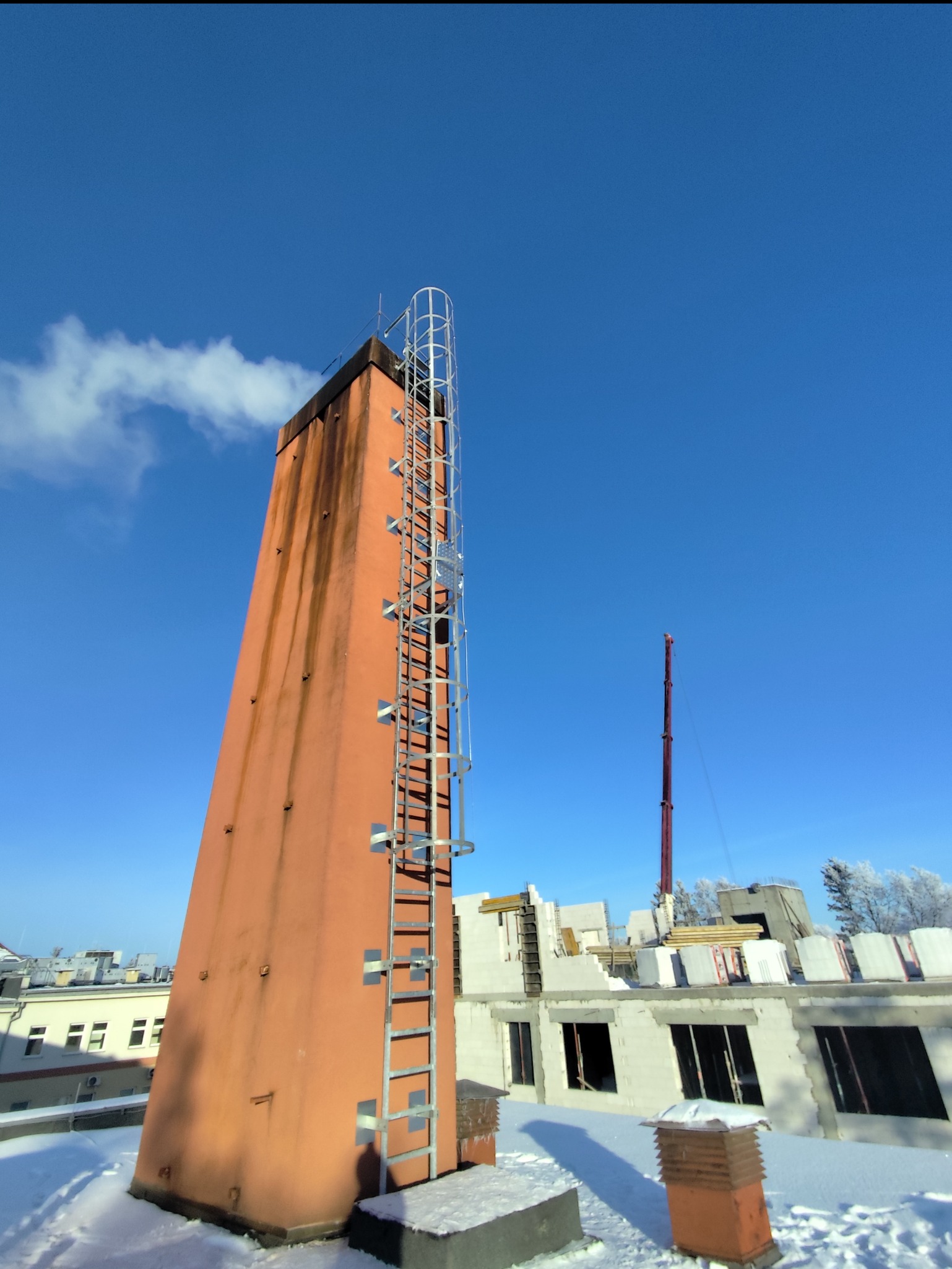Wysoki, pomarańczowy komin z metalową drabiną na dachu budynku w zimowej scenerii, na tle błękitnego nieba. Widoczne ślady zabrudzeń na kominie.