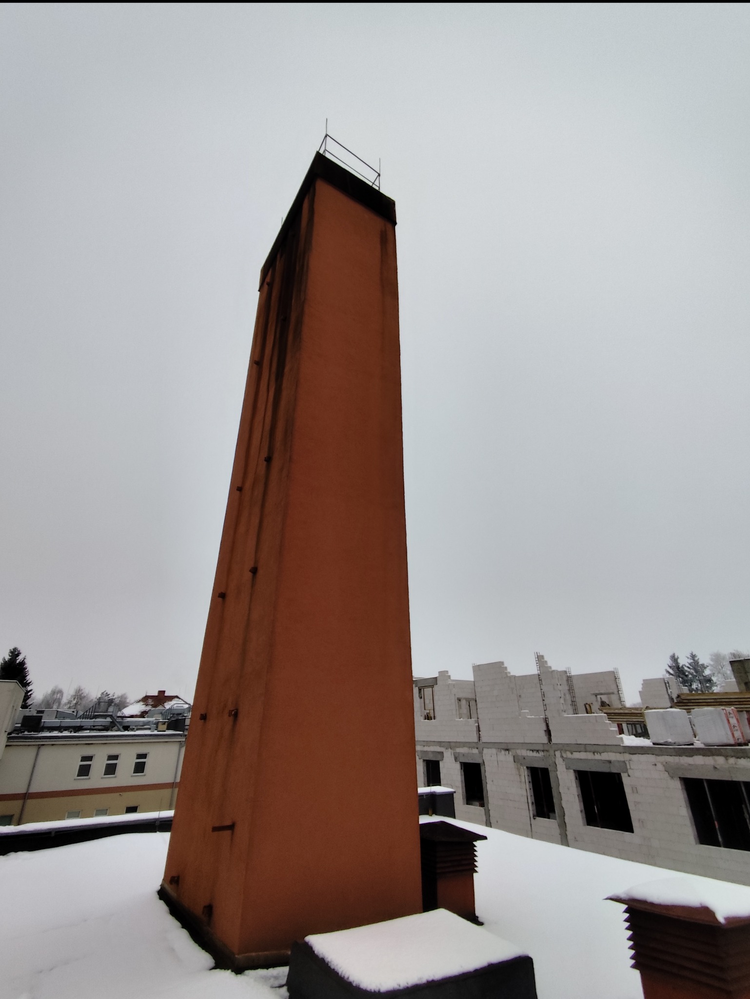 Wysoki, pomarańczowy komin na zasypanym śniegiem dachu, z widocznymi zaciekami. W tle budynek w trakcie budowy i pochmurne niebo.