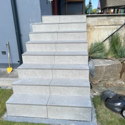 Kacbud - Wykończone schody zewnętrzne z szarych płytek, prowadzące do domu. Widoczny fragment elewacji, trawnik i robot koszący. Ujęcie z dołu, podkreślające solidność wykonania.