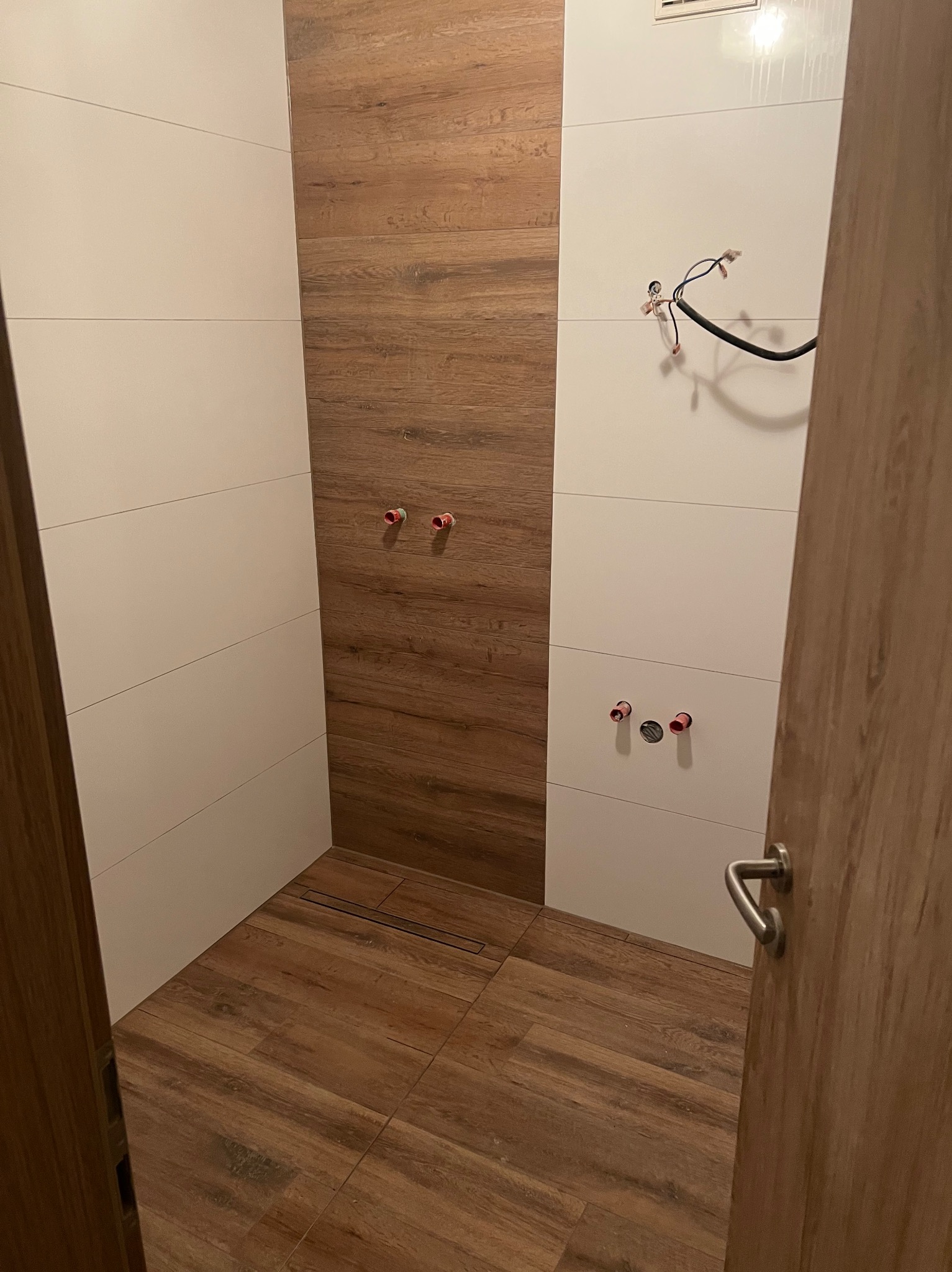 Nowoczesna łazienka w trakcie wykańczania: białe i drewnopodobne płytki, widoczne instalacje wodne i elektryczne, odpływ liniowy w podłodze, otwarte drzwi.