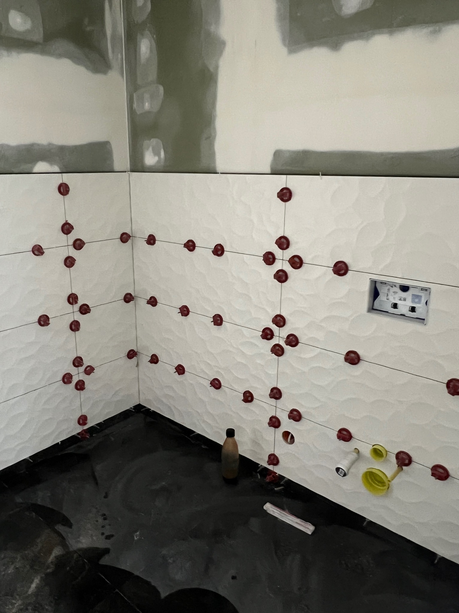 Białe, faliste płytki z czerwonymi krzyżykami dystansowymi, czarna podłoga, etap prac wykończeniowych w łazience. Widoczne narzędzia glazurnicze i niedokończone ściany.