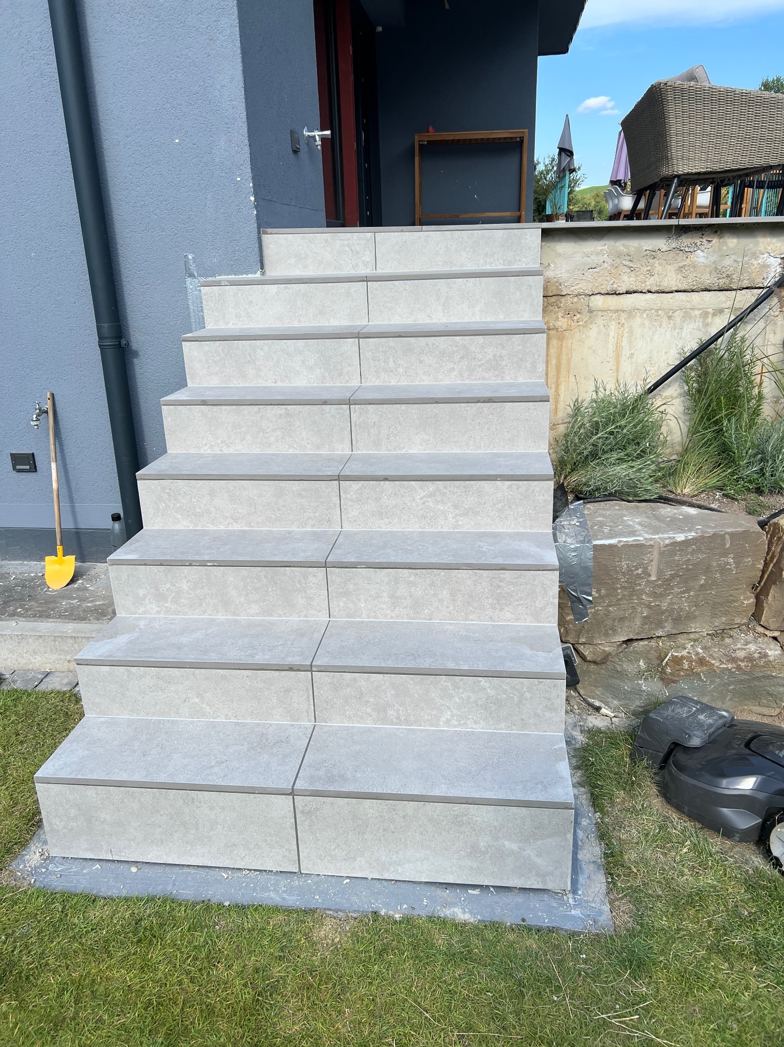 Wykończone schody zewnętrzne z szarych płytek, prowadzące do domu. Widoczny fragment elewacji, trawnik i robot koszący. Ujęcie z dołu, podkreślające solidność wykonania.