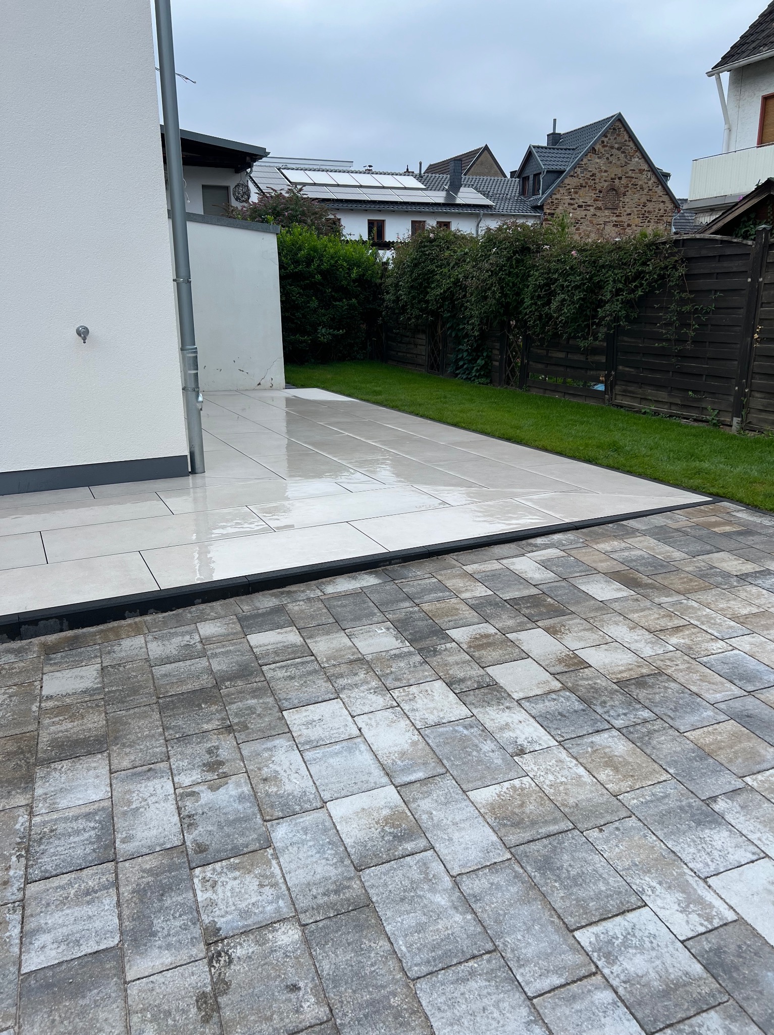 Wyłożony jasnymi płytkami taras przylegający do domu, z widocznym fragmentem kostki brukowej i zieleni w tle. Świeżo po deszczu, mokra nawierzchnia.