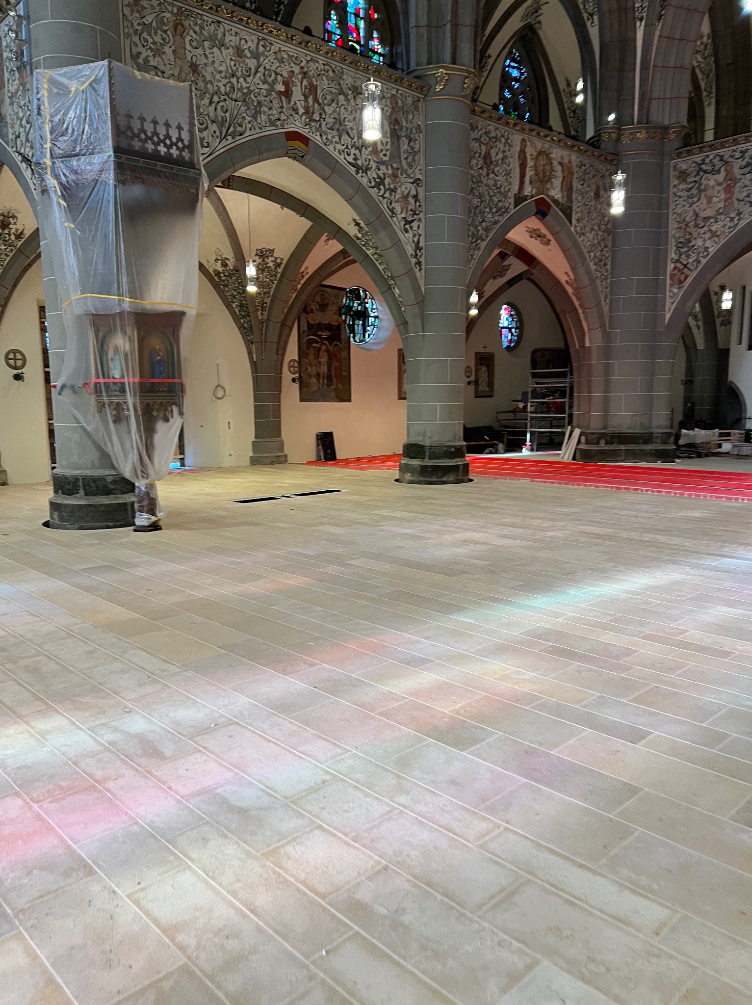 Kamienna posadzka w kościele po renowacji, z widocznymi kolumnami i freskami. Ochronna folia na kolumnie. Światło z witraży barwi podłogę.
