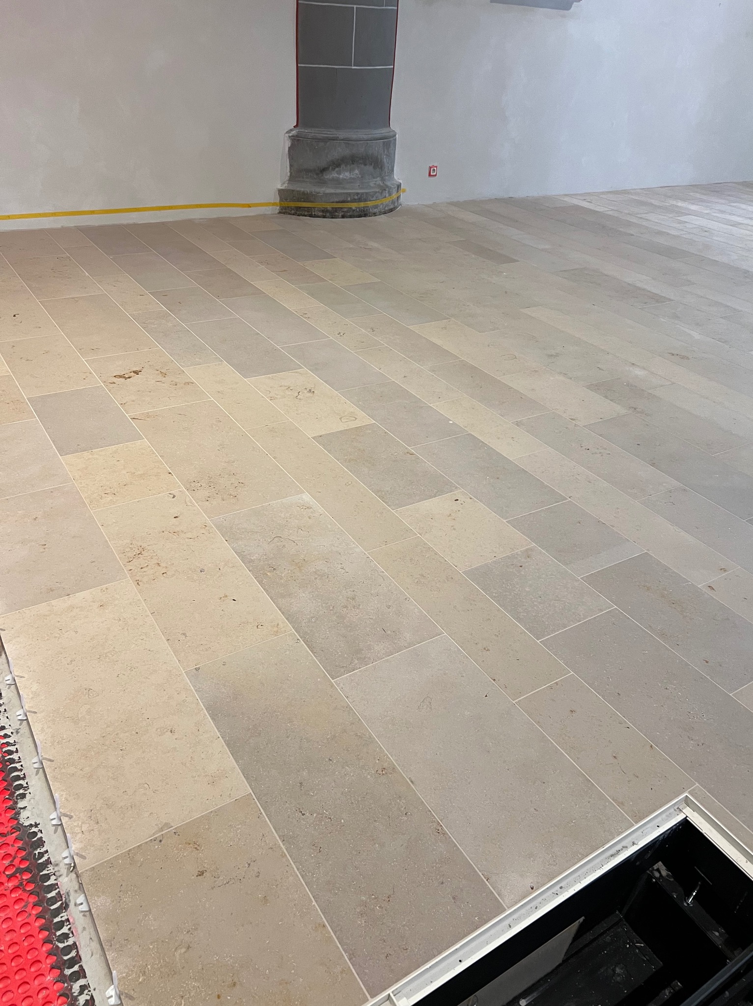 Ułożona, beżowa podłoga z płytek w prostokątnym formacie, widoczna instalacja ogrzewania podłogowego przy ścianie i otwór rewizyjny w rogu pomieszczenia.