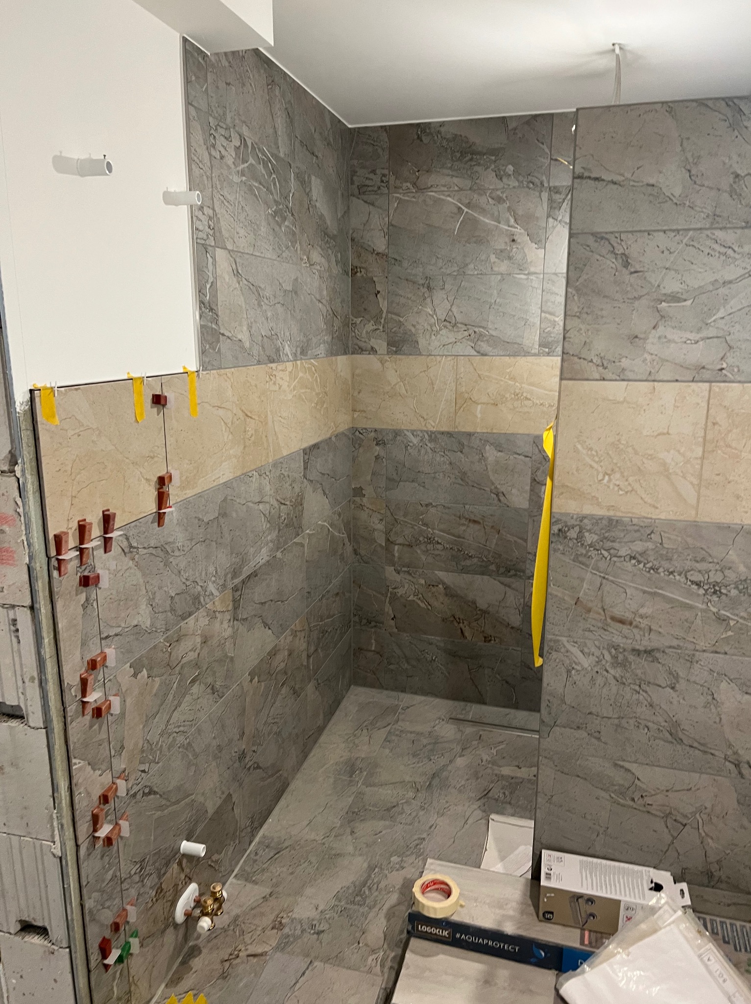 Wnętrze łazienki w trakcie prac glazurniczych. Widoczne płytki imitujące marmur, poziomica, taśma i krzyżyki dystansowe. Częściowo wykończona ściana.