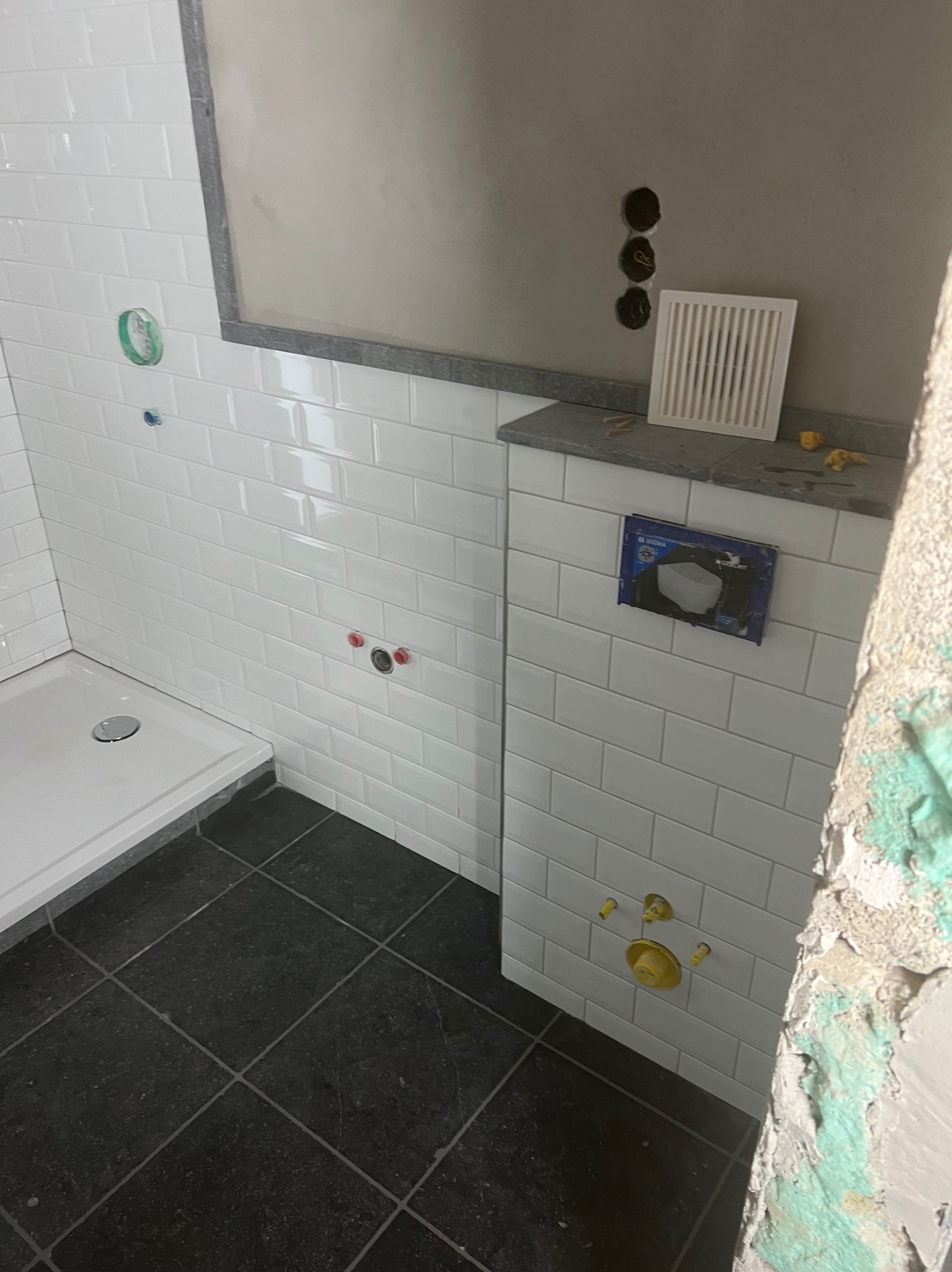 Białe, prostokątne płytki na ścianie łazienki, widoczny fragment kabiny prysznicowej i czarne płytki podłogowe. Częściowo wykończona ściana z instalacją podtynkową.