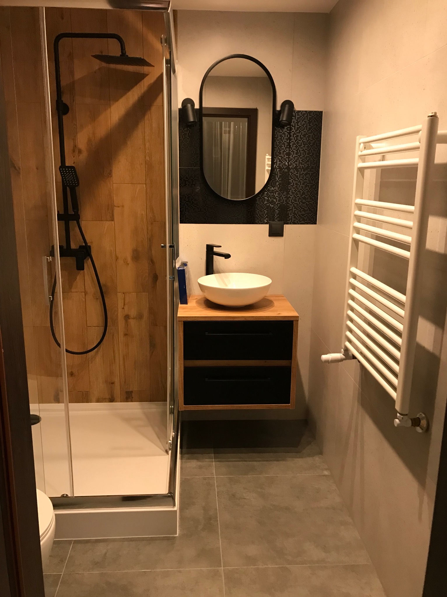 Nowoczesna łazienka z kabiną prysznicową z drewnianymi panelami, umywalką na szafce z czarnymi szufladami i grzejnikiem drabinkowym. Minimalistyczny design.