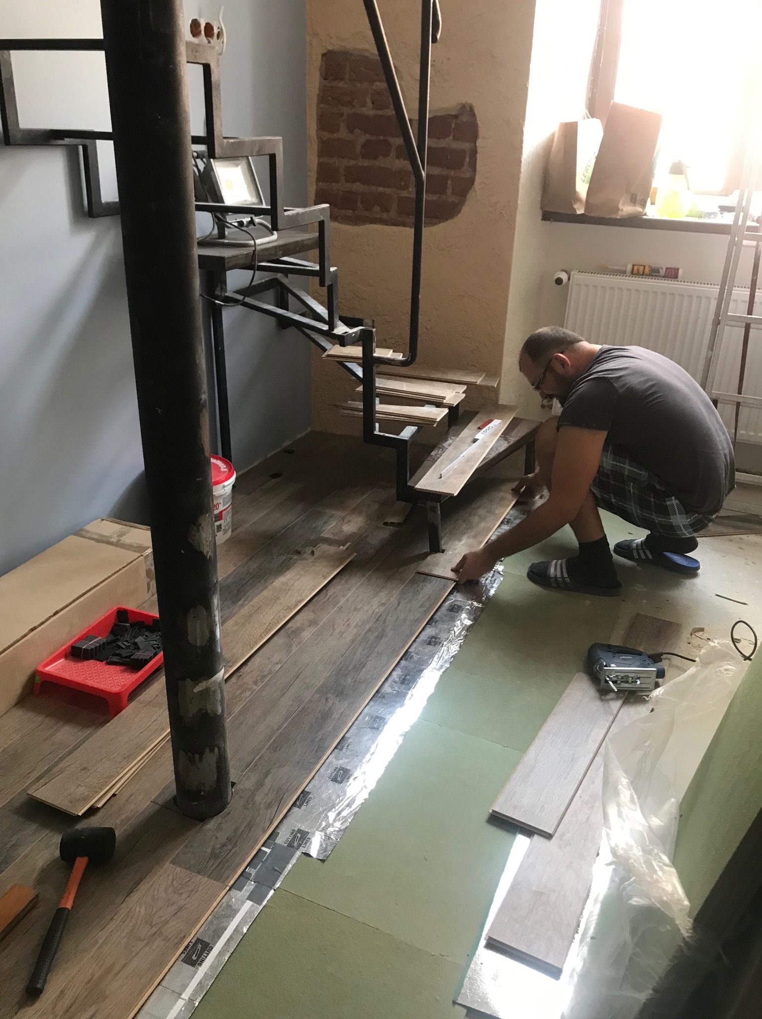 Układanie paneli podłogowych w pomieszczeniu z metalowymi schodami spiralnymi. Widoczny pracownik, narzędzia i materiały. Podłoga z izolacją akustyczną.