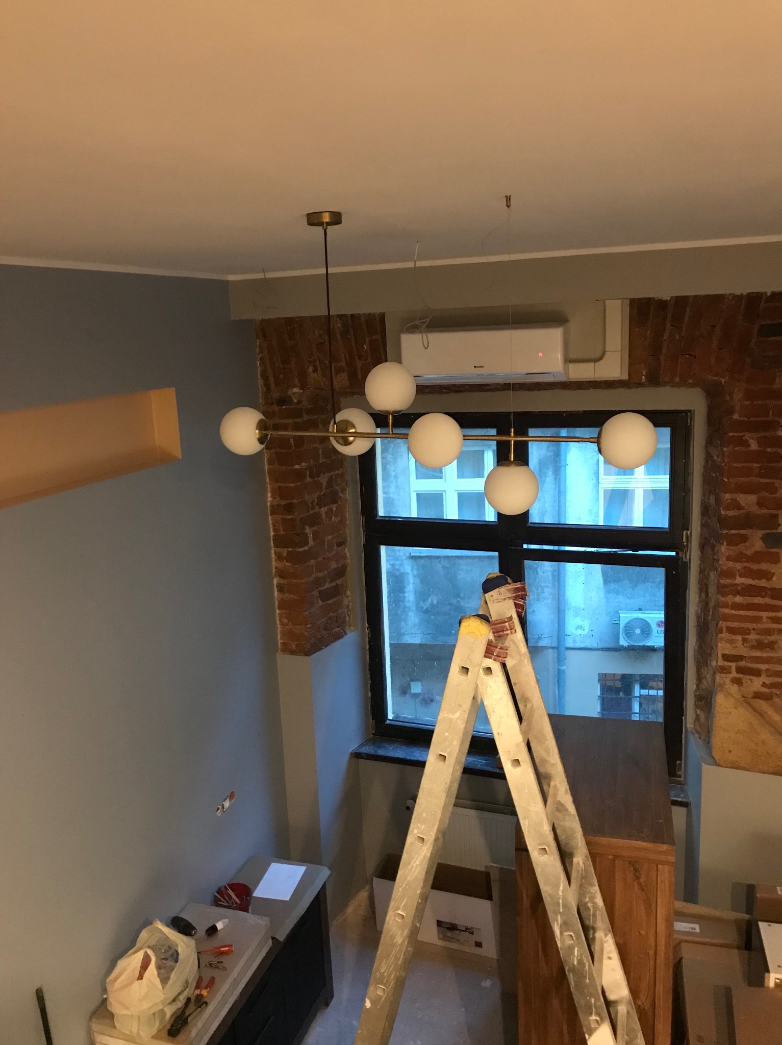 Montaż nowoczesnej lampy kulistej w pokoju z ceglaną ścianą. Widoczna drabina, okno i klimatyzator. Świeże, jasne wnętrze w trakcie wykańczania.