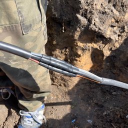 Grzegorz Gierula - Połączenie kabli w ziemi, zabezpieczone termokurczliwymi mufami. Widoczne warstwy kabla i fragment nogi w spodniach moro. Prace ziemne w toku.