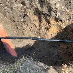 Grzegorz Gierula - Kabel elektryczny w czarnej osłonie ułożony w wykopie ziemnym obok pomarańczowej rury. Widoczne ślady ziemi i trawy.
