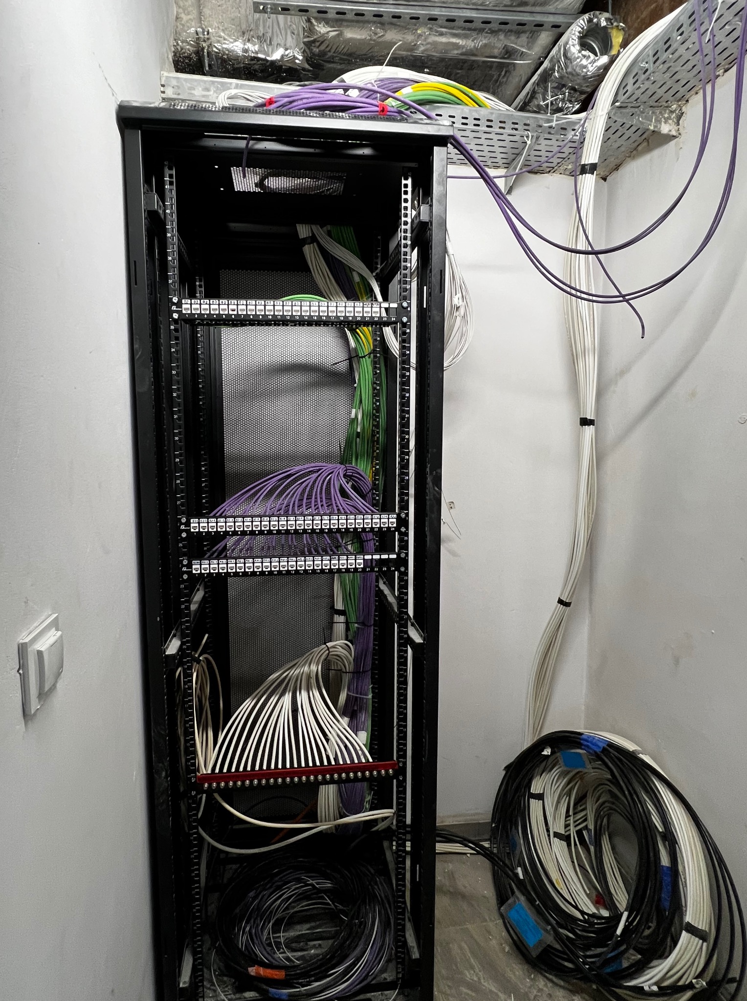 Szafa rack z uporządkowanymi kablami sieciowymi i elektrycznymi w kolorach: fioletowym, zielonym i białym. Kable ułożone w wiązki. Widoczne oznaczenia na panelach.