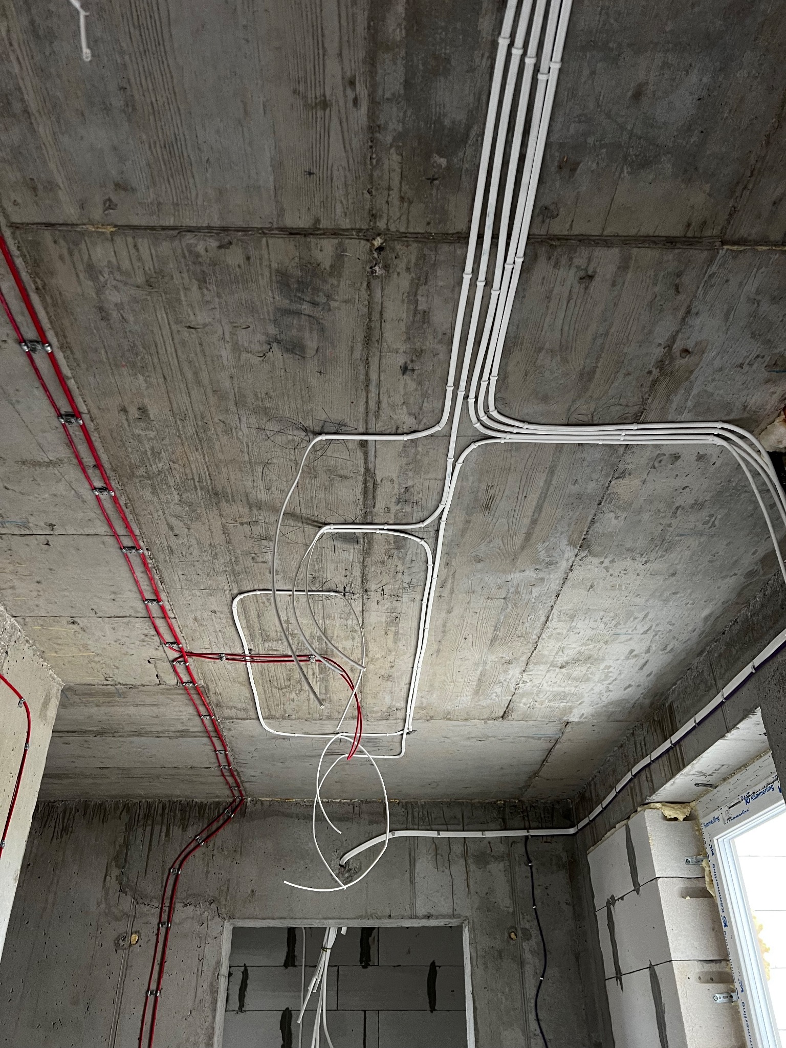 Białe i czerwone kable elektryczne ułożone na surowym, betonowym suficie w trakcie prac instalacyjnych w budynku w stanie deweloperskim. Widoczne uchwyty mocujące.