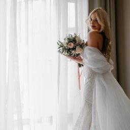 Łukasz Dziopa Fotografia - Panna młoda w koronkowej sukni, z bukietem, pozuje przy oknie z białymi zasłonami. Delikatne światło podkreśla detale sukni i makijaż. Uśmiech skierowany w stronę obiektywu.