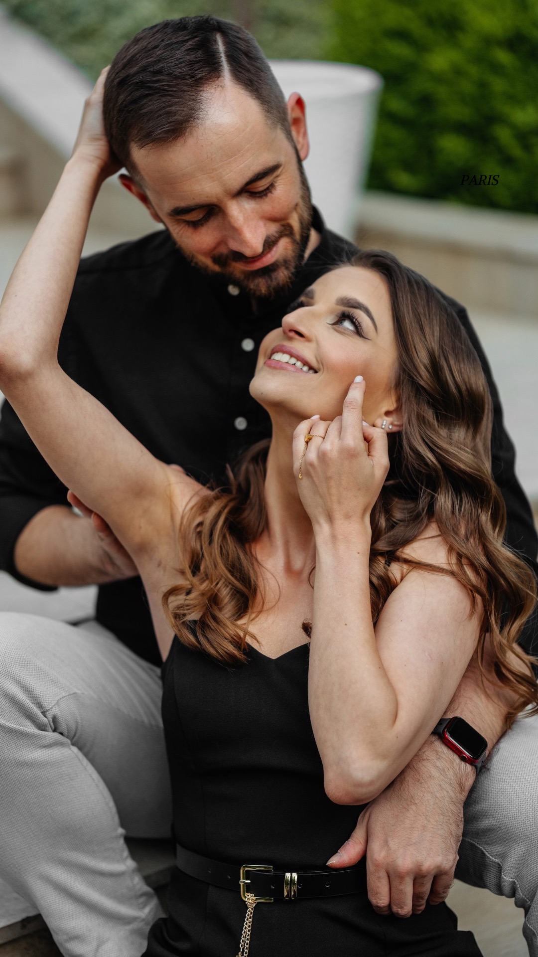 Uśmiechnięta brunetka w czarnej sukience, spogląda w górę, dotykając policzka. Za nią mężczyzna z brodą obejmuje ją, patrząc z czułością. Ujęcie plenerowe, naturalne światło.